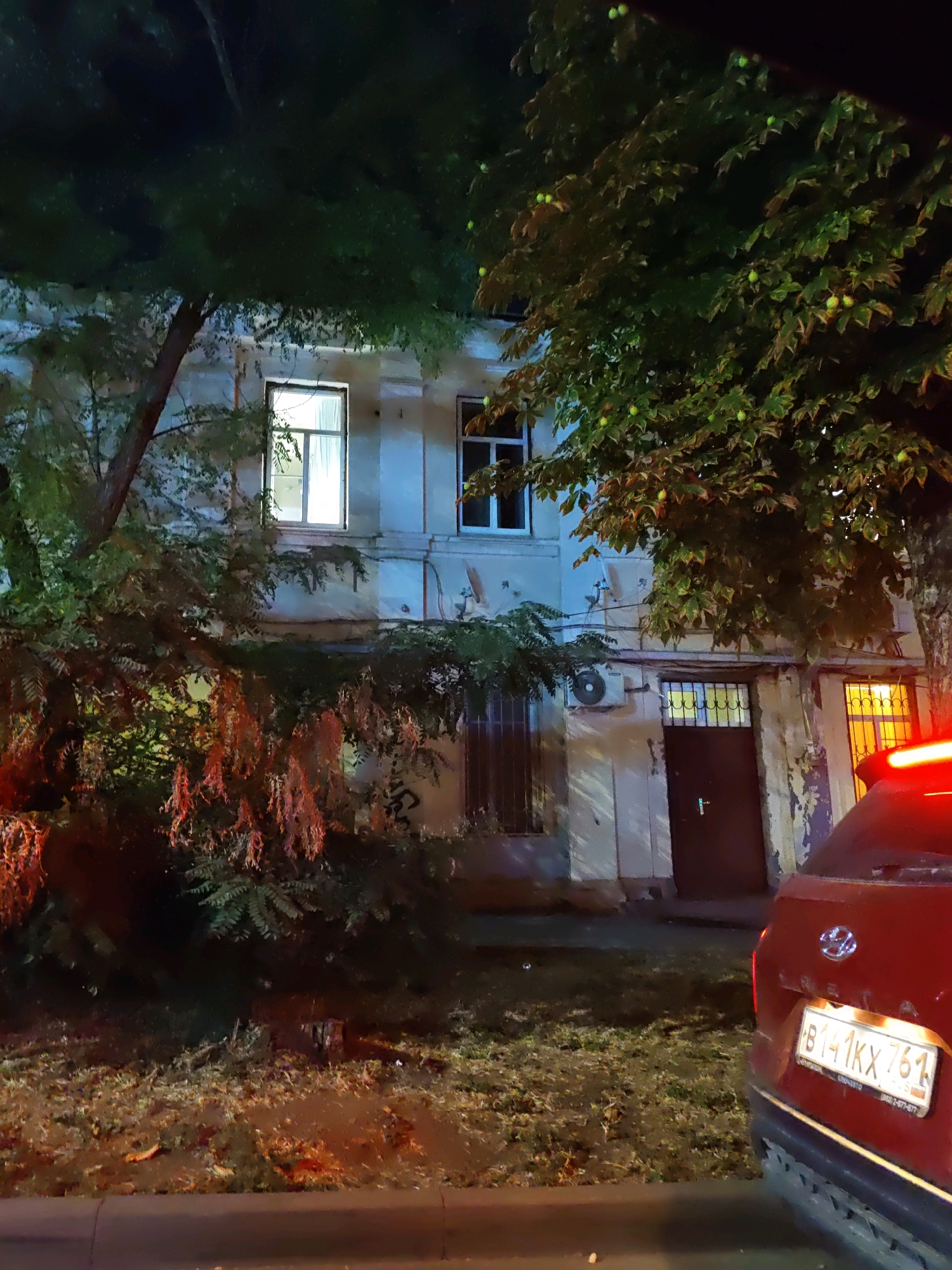 The image shows a nighttime scene in Rostov-on-Don, Russia. The focus is on an old, multi-story building with lit windows. Trees with green leaves and some fruit are in the foreground, partially obscuring the building. A red car is parked in front of the building, with its license plate partially visible, reading "B141KX 761". The street appears to be paved, and there is some vegetation and debris on the ground. The overall atmosphere is dimly lit, suggesting it is late at night or early morning.