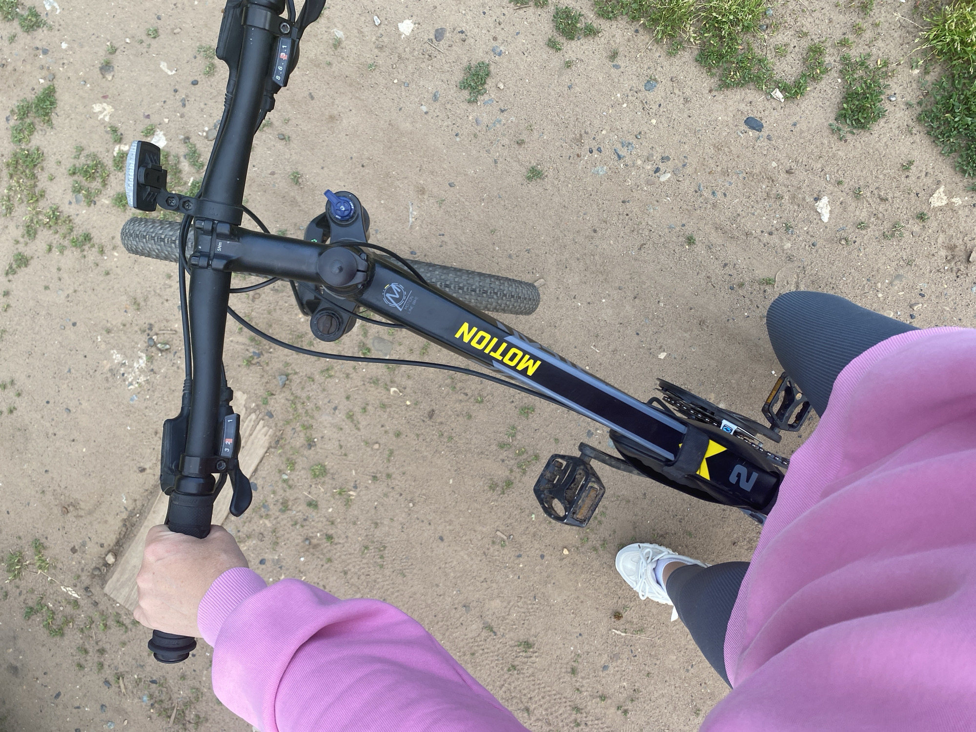 The image shows a first-person perspective of someone on a bicycle. The person is wearing a pink hoodie, dark grey leggings, and white sneakers. Their left hand is on the handlebar, and their right foot is on a pedal. The bicycle is black with yellow and grey accents, and the word "MOTION" is visible on the frame. The front wheel and part of the handlebars are visible. The ground appears to be a dirt or gravel path with scattered grass and small stones. The overall scene suggests an outdoor activity in a casual setting, possibly a park or trail within the city of Chaikovsky, Russia.