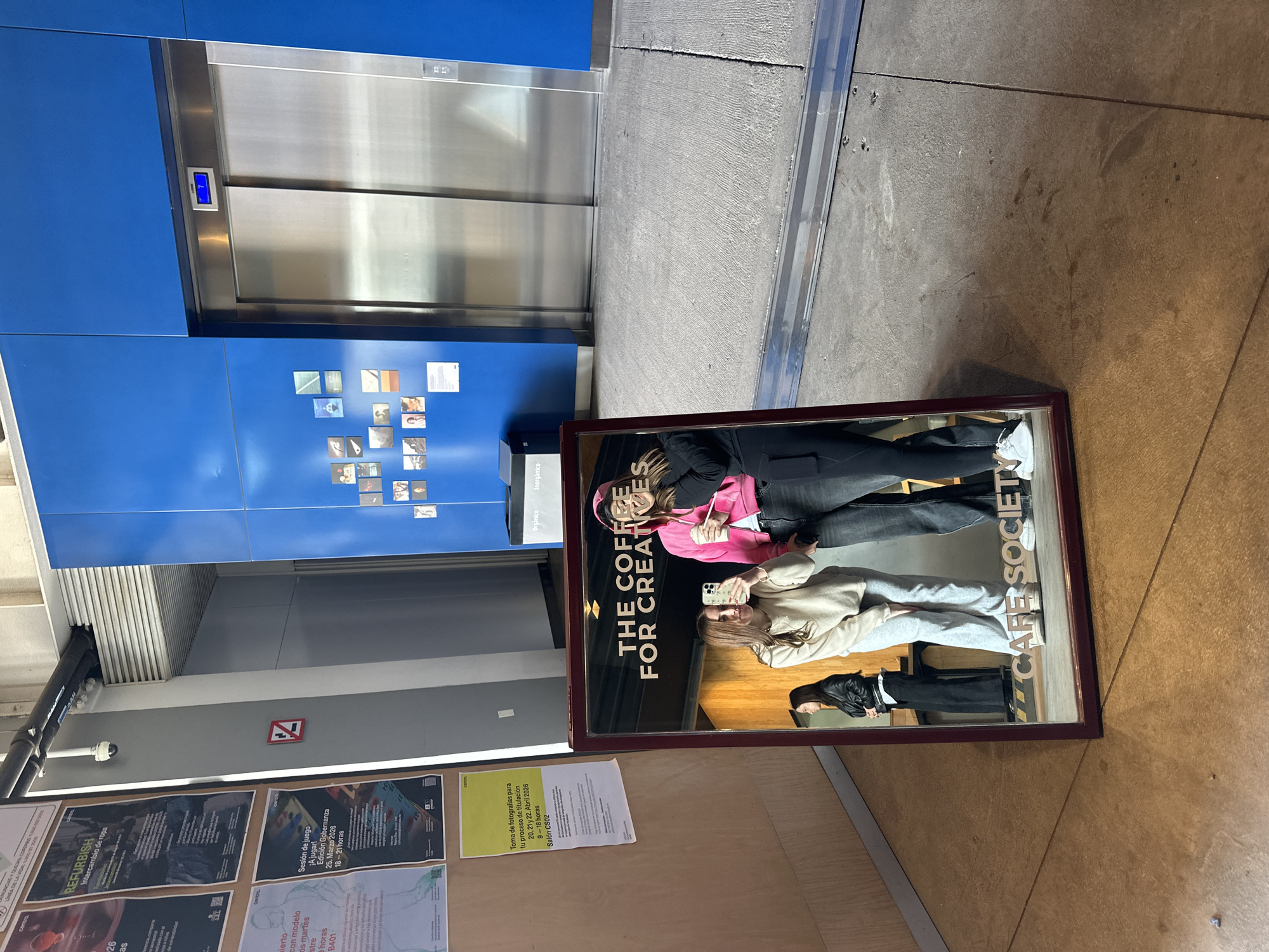 The image shows an indoor setting, likely within a building in Mexico City, Mexico, given the text in Spanish on posters. An elevator is visible with a digital display showing the number "2". The wall next to the elevator is blue with a collection of small, square images. Below this, a waste bin is present. On a wall adjacent to the elevator area, several posters are affixed, including one that mentions "REFURBISH" and another mentioning a "Sesión de juego" on March 25, 2026. A framed sign rests on the floor, reflecting an image of two people. The sign reads "THE COFFEE FOR CREATIVES" and "CAFE SOCIETY". The reflection suggests the two individuals are taking a photo of themselves with the sign. The floor is concrete.