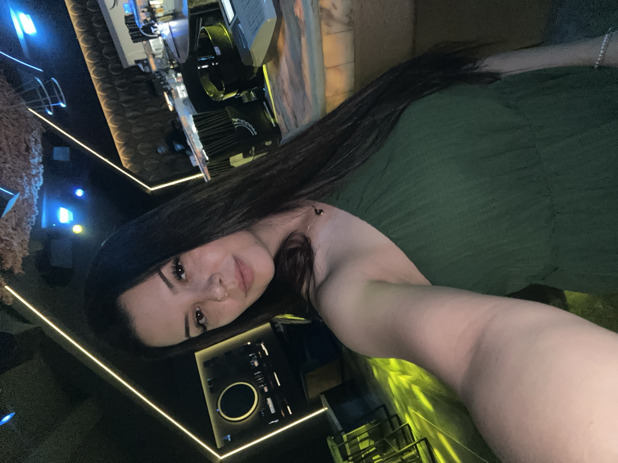 A woman with long dark hair and fair skin with freckles is taking a selfie in what appears to be a dimly lit bar or nightclub. She is wearing a green one-shoulder dress. In the background, there is a bar area with bottles and glasses, as well as some seating areas with yellow lighting. There are also abstract light fixtures on the ceiling. The overall impression is that of a modern, stylish establishment. The city of Sliven, Bulgaria is not mentioned or depicted in this image.