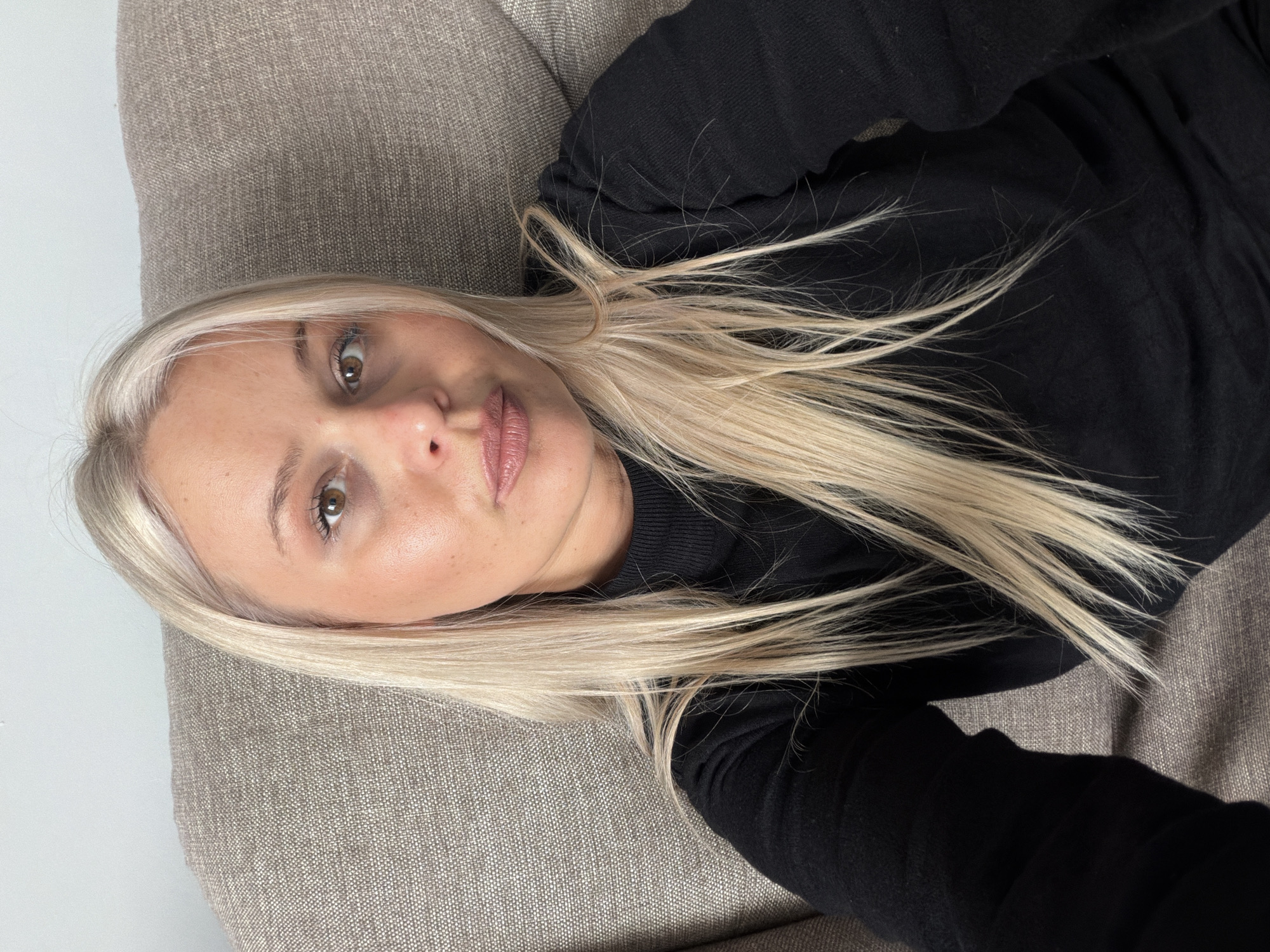 A woman with blonde hair is lying on a gray upholstered couch. She is wearing a black sweater. Her face is visible, and she appears to be looking at the camera. The location is not discernible from the image.