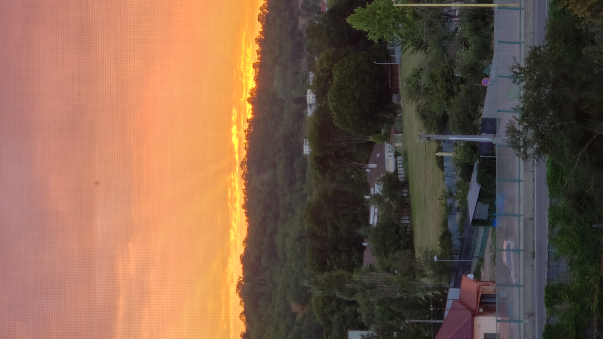 This is a vertical, eye-level shot of a rural or suburban landscape during sunset. The sky is a vibrant gradient of orange and pink, with visible sunbeams breaking through the clouds above a tree-covered hill. Below the hill, several houses with red and white roofs are visible, surrounded by lush green trees. In the foreground, a highway overpass with a fence runs horizontally across the image, with cars and greenery visible on and around it. The image appears to have been taken through a screen or window, as a grid pattern is visible overlaying the scene. The location is identified as being in Prešov, Slovakia, based on provided metadata. There are no people or discernible interactions visible in the scene.