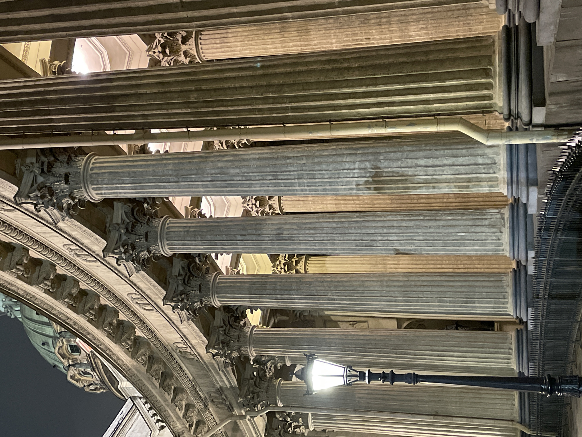 This appears to be an upward-angled shot of the colonnade of the Kazan Cathedral in Saint Petersburg, Russia. The image captures multiple rows of tall, gray stone columns with fluted surfaces. The columns feature ornate Corinthian capitals. There is a black lamppost in the foreground. The sky is dark, suggesting it is nighttime. The perspective creates a strong sense of vertical lines and repetition.