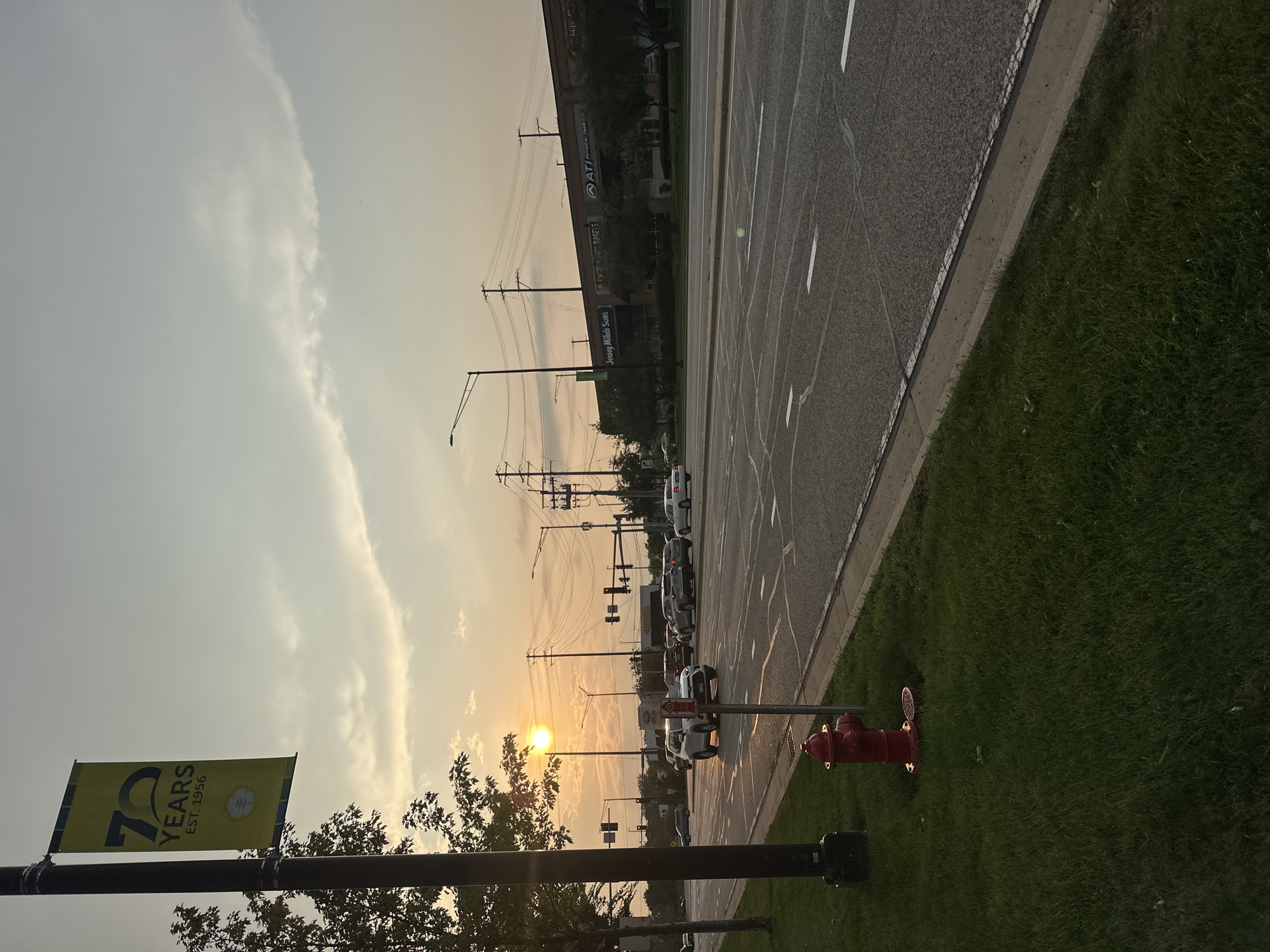 This image depicts a street scene in Schaumburg, Illinois, at dusk. The sun is low in the sky, casting a warm glow and creating long shadows. A banner on a lamppost celebrates "70 YEARS EST. 1956." Cars are driving on a multi-lane road, and a red fire hydrant is visible on the grassy median. In the background, a row of businesses, including "Jersey Mike's Subs" and "ATI Physical Therapy," are illuminated by streetlights. The sky is partly cloudy, with hints of orange and yellow from the setting sun. The overall atmosphere is serene and suburban.