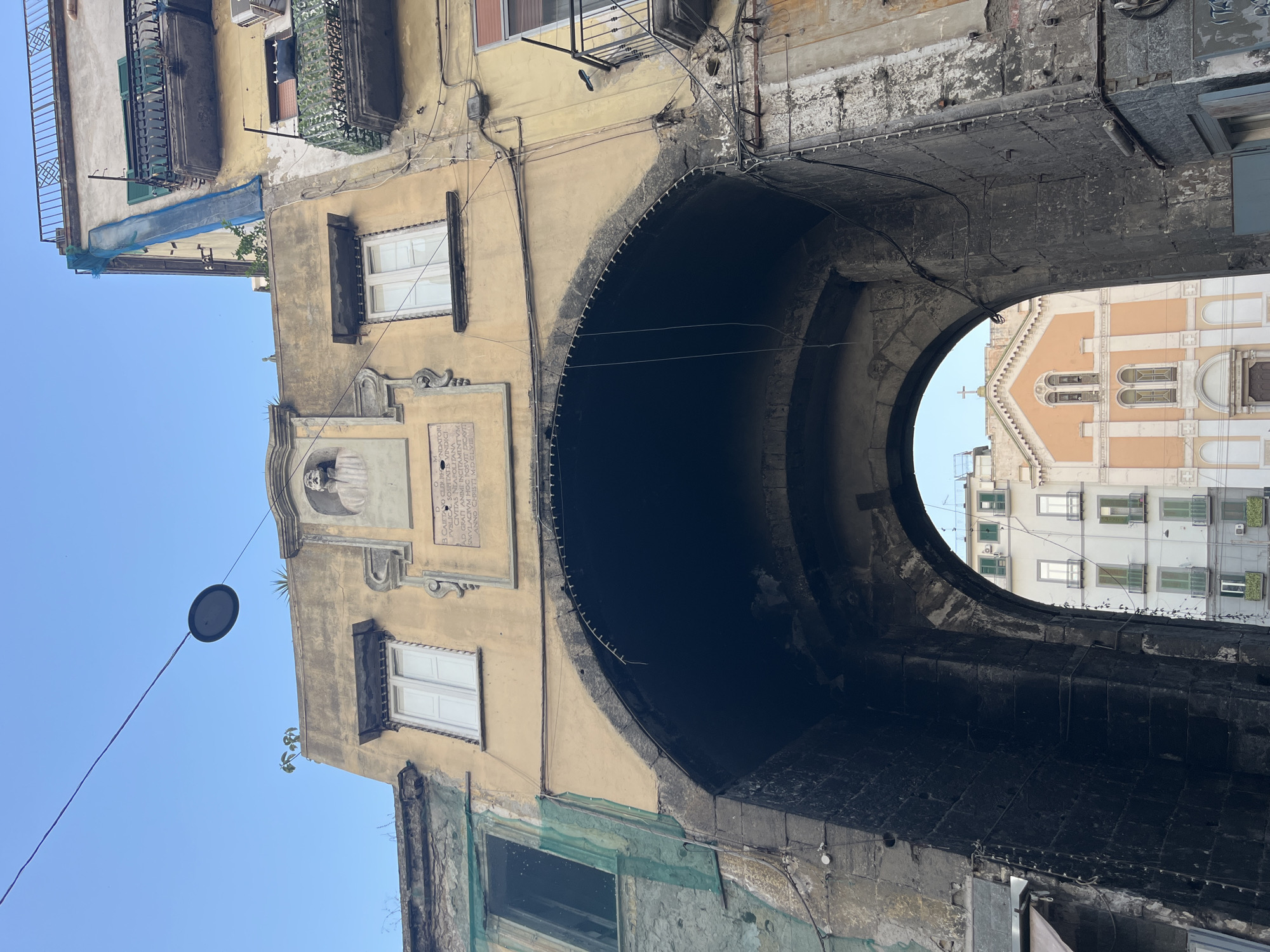The image captures a view looking up through a stone archway in Naples, Italy. The archway is part of an old building with weathered yellow stucco. Above the arch, a plaque with an inscription and a bust of a man are visible. To the sides of the plaque are two windows with white shutters. The building has balconies with iron railings and some are covered with netting or camouflage material, suggesting ongoing construction or renovation. Electrical wires are strung across the facade.
Through the arch, a church with an orange facade and a cross on its roof is visible, along with other buildings with green shutters. The sky above is clear blue. The overall impression is of a historical urban setting with signs of both age and recent activity. The specific location is likely an ancient gateway or entrance within the historic center of Naples.