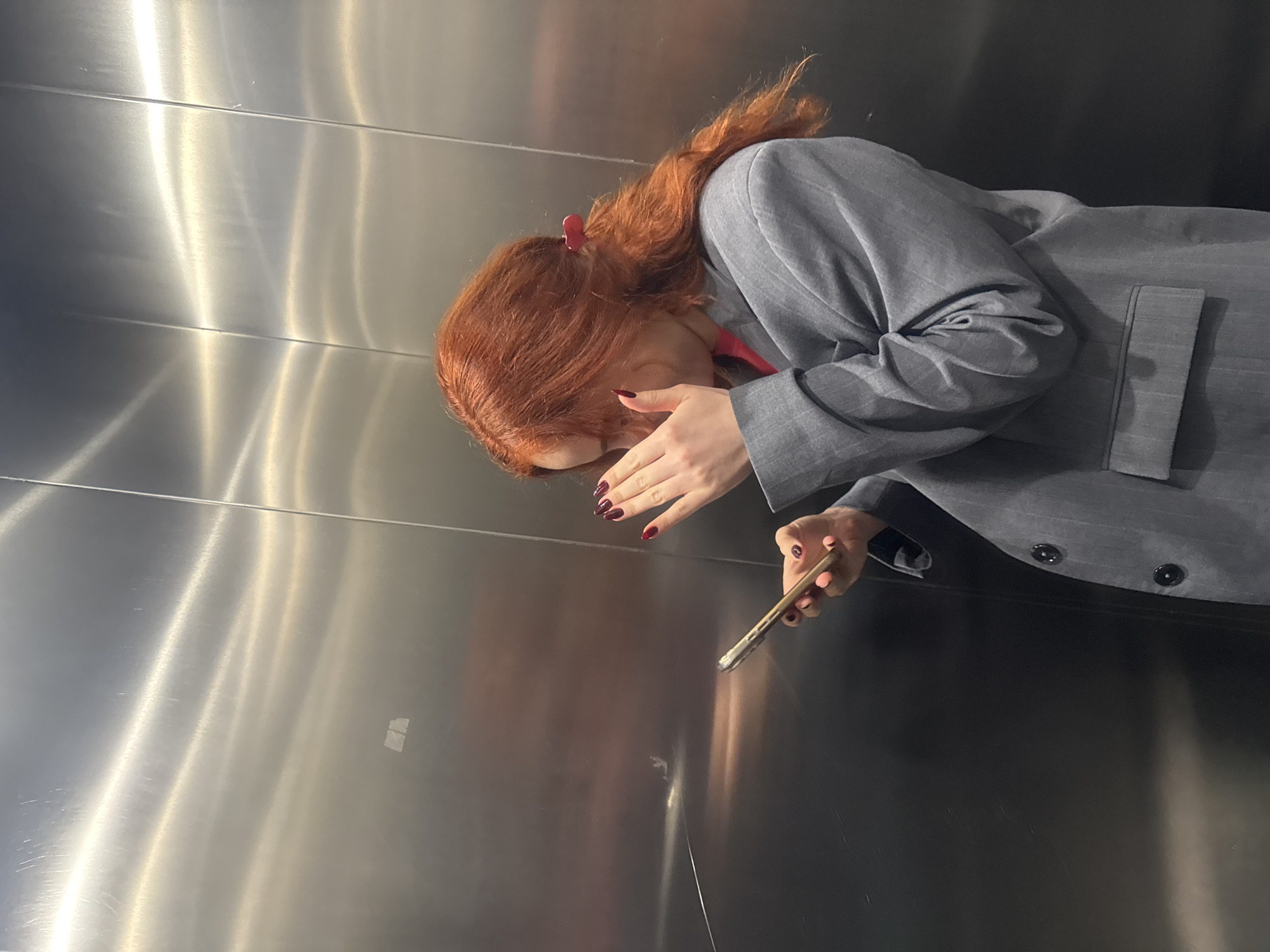 A woman with long red hair, partially pulled back with a red hair clip, is leaning against a stainless steel or silver colored paneled wall that appears to be inside an elevator in the city of Tbilisi, Georgia. She is wearing a gray pinstriped blazer. Her left hand is held up to her face, covering part of it, and she is holding a cell phone in her right hand. She has dark red nail polish on both hands. The background is dark, suggesting the elevator is not well-lit.