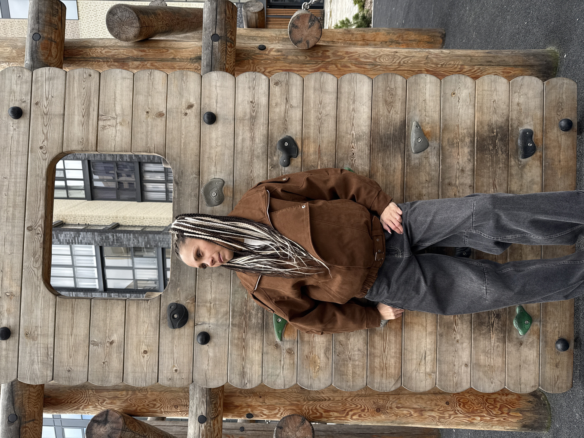 The image depicts a person with braided hair wearing a brown jacket and gray pants lying on a wooden climbing wall structure. The wall is composed of vertical wooden planks, featuring various climbing holds in different shapes and colors attached to it. Parts of a building with multiple windows are visible in the background through an opening in the wooden wall. The environment suggests an urban playground or recreational area.