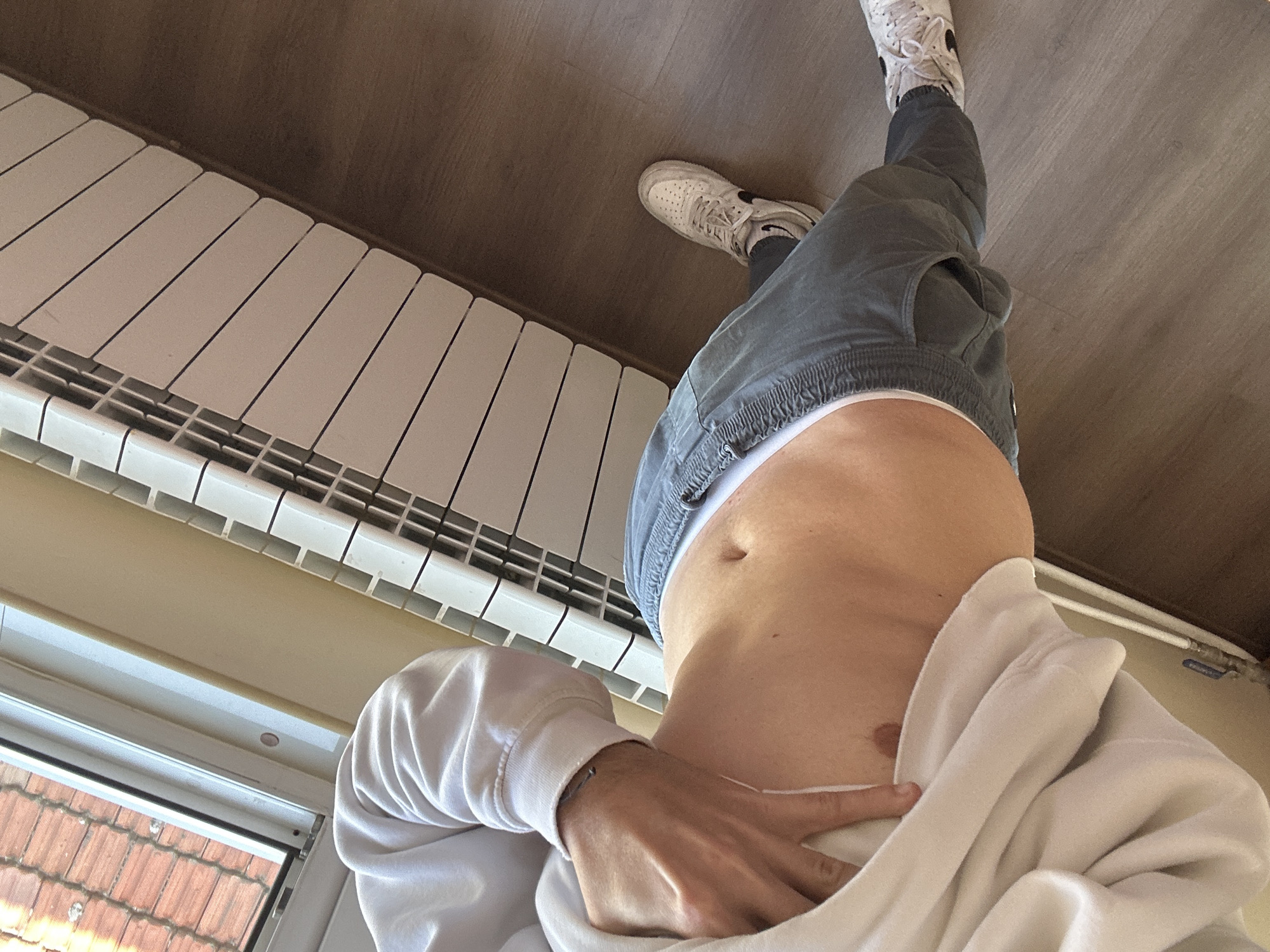 The image captures a person from a low-angle perspective, focusing on their torso, right arm, and lower body. The person is wearing a white hoodie, grey cargo-style pants with an elasticated waist, and white sneakers, which appear to be Nike Air Force 1s with a black swoosh logo visible on one shoe. A white underwear waistband is visible above the pants. The person's right hand is casually holding up the bottom edge of the hoodie, revealing their abdomen.
The setting is indoors, with a prominent white radiator running along a wall. The floor or a wooden panel above the radiator is visible. In the lower left, a window provides a glimpse of a terracotta-tiled roof outside, indicating daytime conditions. The overall scene suggests a casual environment. There is no other discernible text apart from the Nike logo on the sneaker. The context provided indicates the location is Plovdiv, Bulgaria.