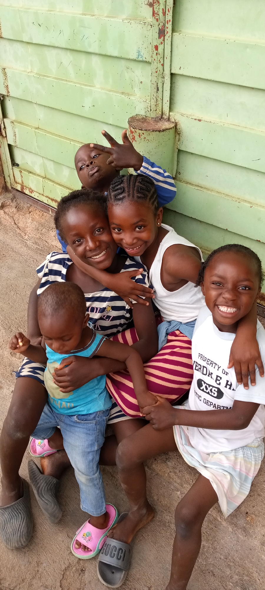 A group of five children are huddled together in an outdoor setting. The backdrop is a light green, horizontally corrugated metal structure.
The children are dark-skinned and are varying in age. The youngest child, a toddler, is wearing a blue t-shirt and jeans, seated on the lap of an older child wearing a striped shirt. There is a child wearing a white tank top. Another child in the front right is wearing a white t-shirt with "PROPERTY OF VERDKE HALL" printed on it. Another child is standing behind.
The group is sitting on a concrete ground. Several pairs of slides are visible, indicating the children are likely barefoot.
The setting appears to be a casual, possibly residential, environment.