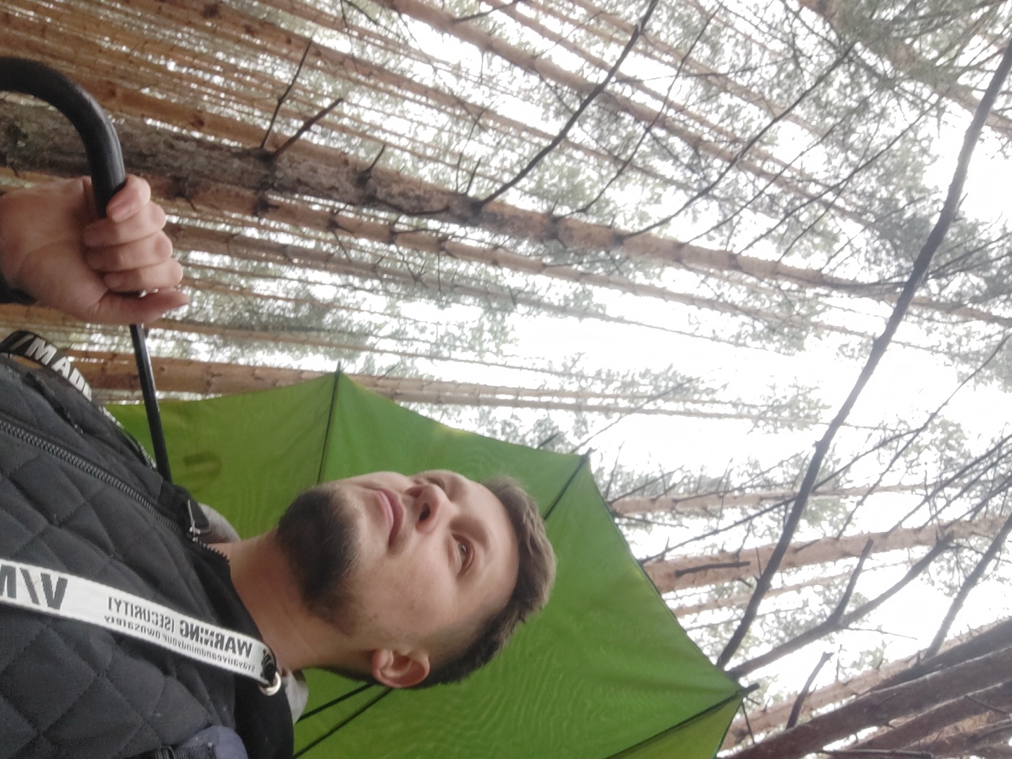 This image, likely rotated 90 degrees counter-clockwise, depicts a man outdoors in a forest, possibly in Chaikovsky, Russia, on an overcast day. The main subject is a man with short brown hair and a beard, seen from the chest up, looking upwards and slightly to his right with a contemplative expression. He is wearing a black quilted jacket or vest over a dark top. Around his neck, a white lanyard is visible, displaying reversed text that clearly reads "WARNING SECURITY".
His hand, visible in the upper left of the frame, grasps the curved handle of a black umbrella or a similar pole. A large green umbrella or tarp is partially visible behind and above the man, providing shelter. The background features a dense canopy of tall, slender trees, likely pines, with numerous branches silhouetted against a bright, uniform overcast sky, suggesting diffuse light and no direct sunlight. The scene conveys a sense of being immersed in nature, perhaps taking a break or seeking shelter from potential rain.
