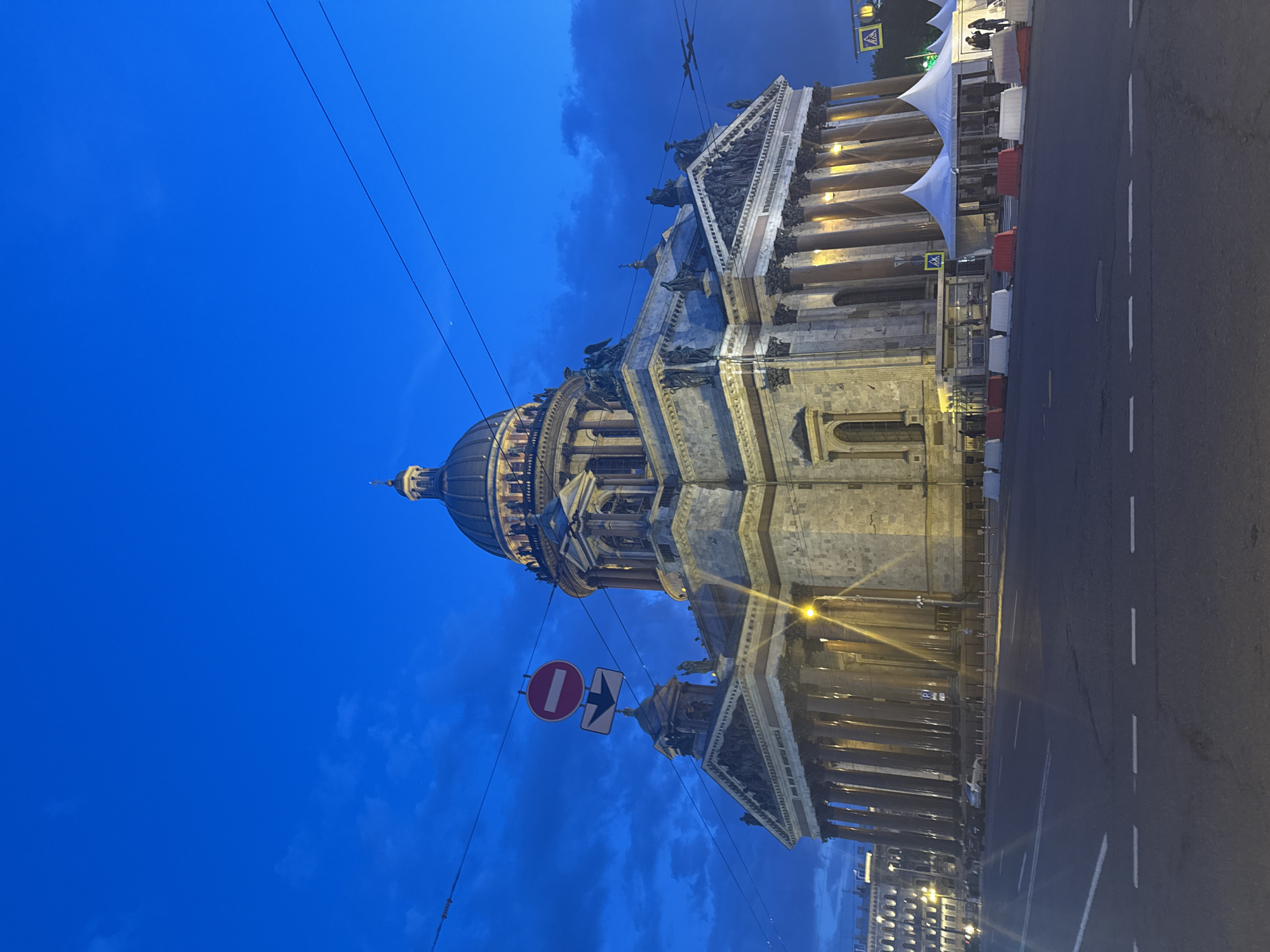 The image captures a grand architectural landmark, Saint Isaac's Cathedral in Saint Petersburg, Russia, at what appears to be twilight or early night. The building, illuminated by artificial lights, dominates the scene with its classical design, featuring numerous massive columns, detailed pediments adorned with sculptures, and a prominent golden dome topped with a lantern and spire.
The sky is a deep, dark blue, indicating the late hours, with some visible clouds adding texture. Below the cathedral, a wide asphalt road is visible, marked with white dashed lines. Overhead wires crisscross the upper portion of the sky.
On the right side of the image (as if rotated to correct orientation), alongside the cathedral, there are several white pyramid-shaped tents or canopies, possibly temporary structures for an event or market. In front of these tents, red and white concrete barriers line the roadside. Small figures, likely people, can be discerned near these structures and on the steps of the cathedral.
Several traffic signs are visible: a circular red "no entry" sign with a white horizontal bar, a rectangular white sign with a black arrow pointing right, and a yellow diamond-shaped sign with a black pedestrian crossing symbol. Streetlights emit a warm glow, further illuminating parts of the street and the cathedral's facade. The overall mood is serene and impressive, highlighting the cathedral's majestic presence against the evening sky.