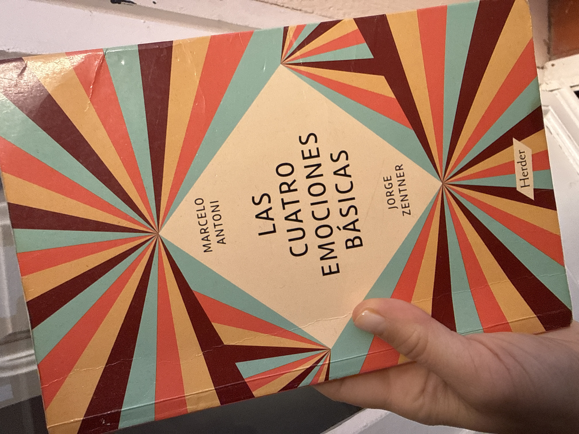 A person holds a book titled "Las Cuatro Emociones Basicas" by Marcelo Antoni and Jorge Zentner, published by Herder. The book has a cover design featuring radiating stripes in teal, orange, yellow, and burgundy. The background is a white surface, likely a shelf or table, and a hint of a door frame is visible in the upper left corner. The location is not discernible from the visual data to determine its presence in Barcelona, Spain.