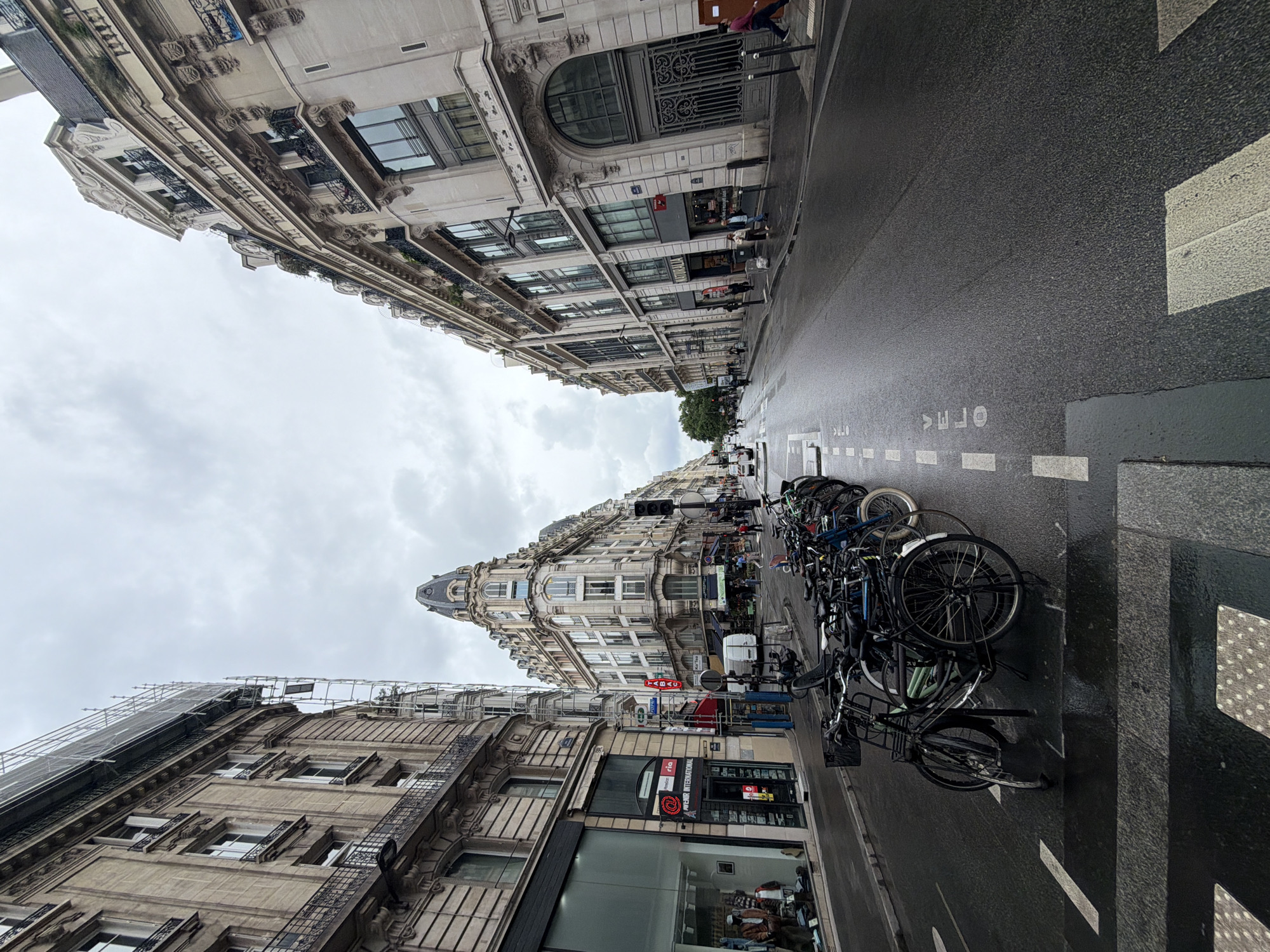 The image captures a street scene in Paris, France, characterized by tall, ornate buildings lining both sides of a wet, asphalt street. The perspective is from ground level, tilted slightly to emphasize the height of the buildings. The sky is overcast. On the right side of the street, near the foreground, numerous bicycles are parked, creating a dense cluster. A crosswalk with white markings is visible in the lower right corner. The street's surface is dark, reflecting the overcast sky and suggesting recent rainfall. Some pedestrians can be seen further down the street, interacting near storefronts. The architectural style of the buildings is consistent with Parisian architecture, featuring detailed facades, balconies, and intricate ornamentation. One building features a distinctive corner tower with a dome-shaped roof. Shop signs and advertisements are visible, confirming the commercial nature of the area.