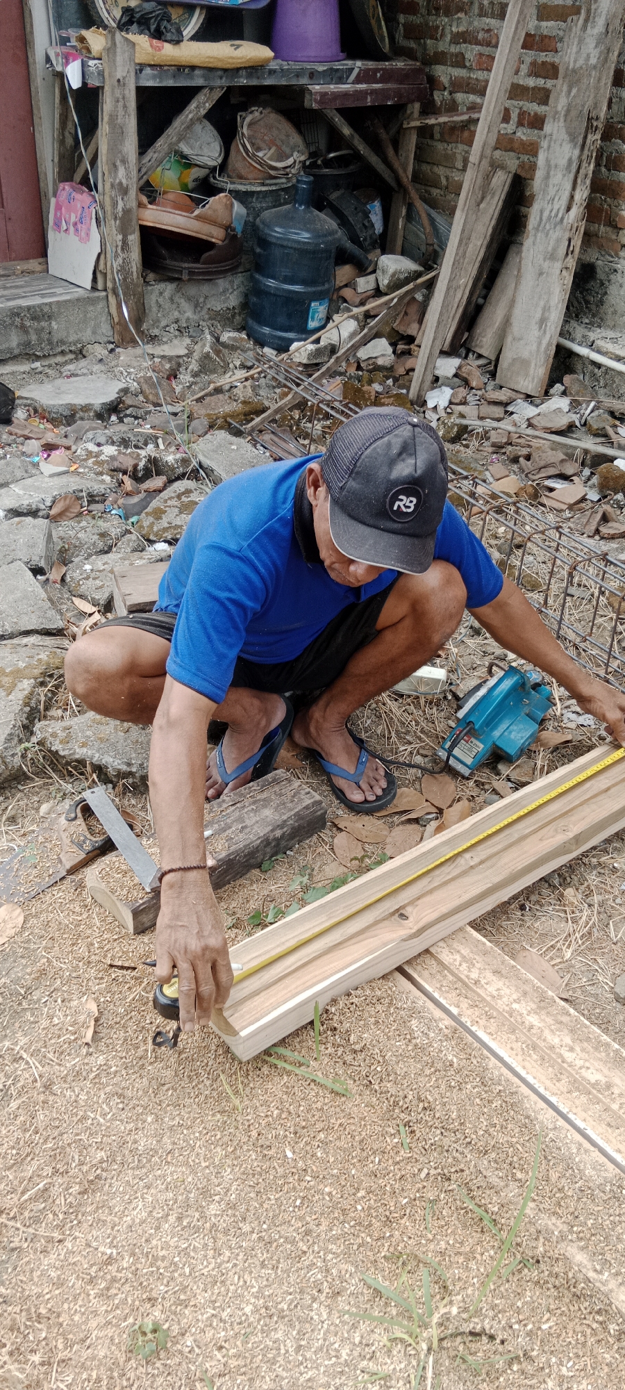 In Pelang, Indonesia, a man in a blue shirt and black shorts is squatting on the ground, measuring a piece of wood. He wears a black baseball cap with an "RB" logo and blue flip-flops. In front of him, a yellow measuring tape stretches across the wooden plank, which rests on sawhorses. To his left, a saw lies on a wooden block. To his right, a turquoise power tool, possibly a sander, sits on the ground. The environment appears to be an outdoor workspace littered with debris, including rocks, broken bricks, and wood scraps, indicative of construction or renovation activity. In the background, a brick wall and various discarded items, such as buckets and large water containers, are visible.