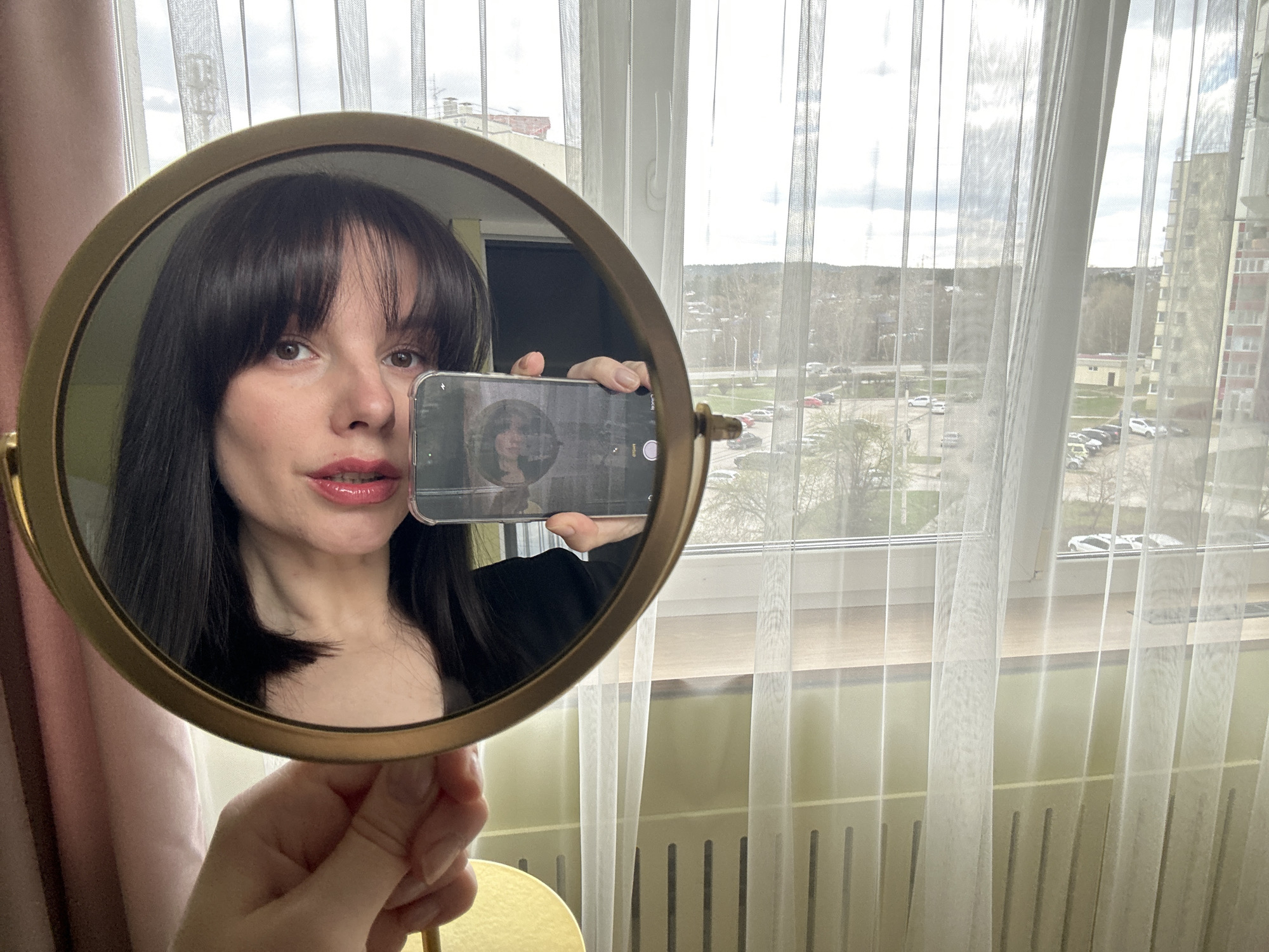 A woman is holding a round, gold-framed mirror in her left hand, using it to reflect her image while simultaneously capturing it on a smartphone held in her right hand. The reflection shows her face with dark hair and bangs, as well as a hint of her dark-colored clothing. The phone's screen displays a grayscale image of the woman. The background features a window covered with white, sheer curtains. Outside the window, a cityscape is visible, including buildings, trees, and parked cars. Based on the architectural style and urban layout, the location is identified as Chaikovsky, Russia.