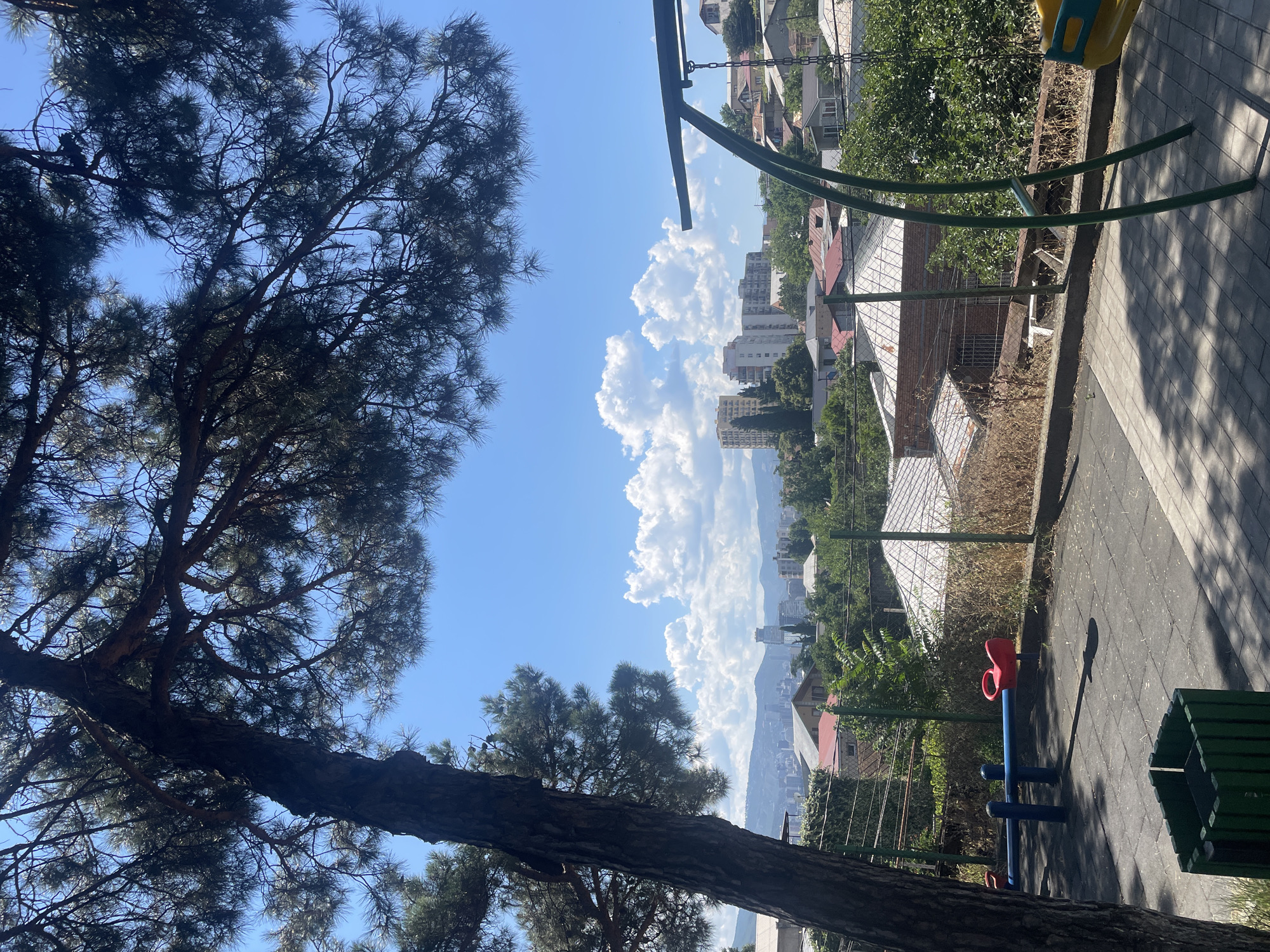 This is a vertical, medium-shot, eye-level, outdoor photograph taken on a sunny day with a clear blue sky dotted with puffy white clouds. The foreground features a playground with a seesaw, a swing set, and a green trash receptacle. The playground surface is made of gray rubber tiles with a checkered pattern. A large pine tree with dark green needles dominates the left side of the frame, its branches extending across the top and obscuring parts of the sky.
Beyond the playground, a chain-link fence separates the area from a slope covered in dry grass and vegetation. Further in the background, the roofs of houses with red and white tiles are visible, nestled among lush green trees. In the middle ground, apartment buildings of varying heights and architectural styles, some with balconies and white or yellow facades, rise above the residential structures.
The distant background showcases a cityscape with more buildings and a hazy mountain range forming the horizon. The overall impression is of a residential area in Tbilisi, Georgia, situated on a hillside overlooking the city. The lighting suggests it is midday or early afternoon, with distinct shadows cast by the tree and playground equipment on the ground. There are no people or animals present in the image.