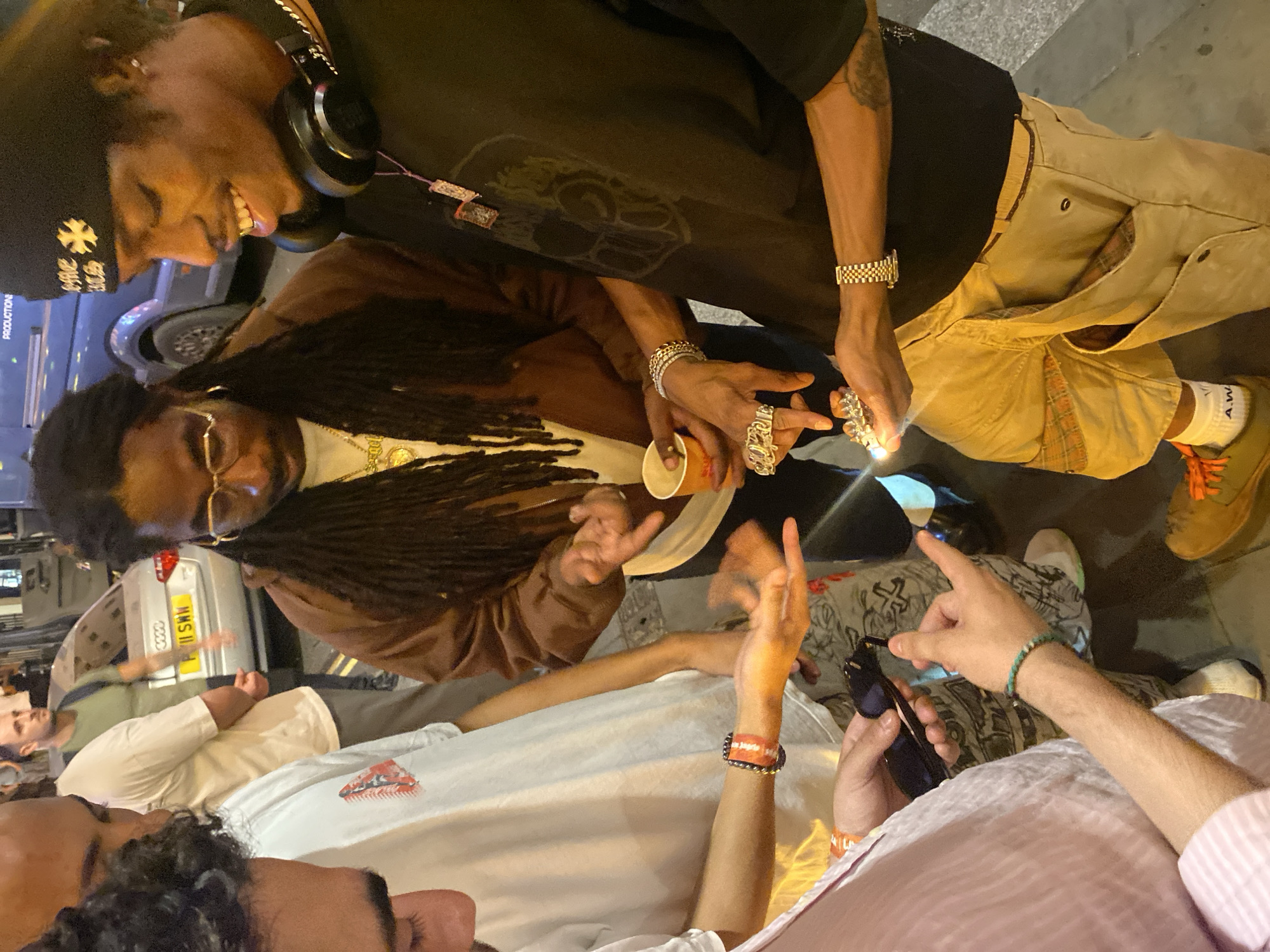 A group of people are gathered outdoors at night in what appears to be a street location in London, United Kingdom. One person with dreadlocks is wearing a brown jacket, while another nearby person is in a dark colored T-shirt and a cap with headphones. There is a silver Audi car in the background with the license plate "MNS 11". Some individuals are holding objects such as a cup and a phone. One of the people is holding a light, perhaps a lighter. The other people appear to be interacting with each other. The overall environment is urban, with street lighting visible.