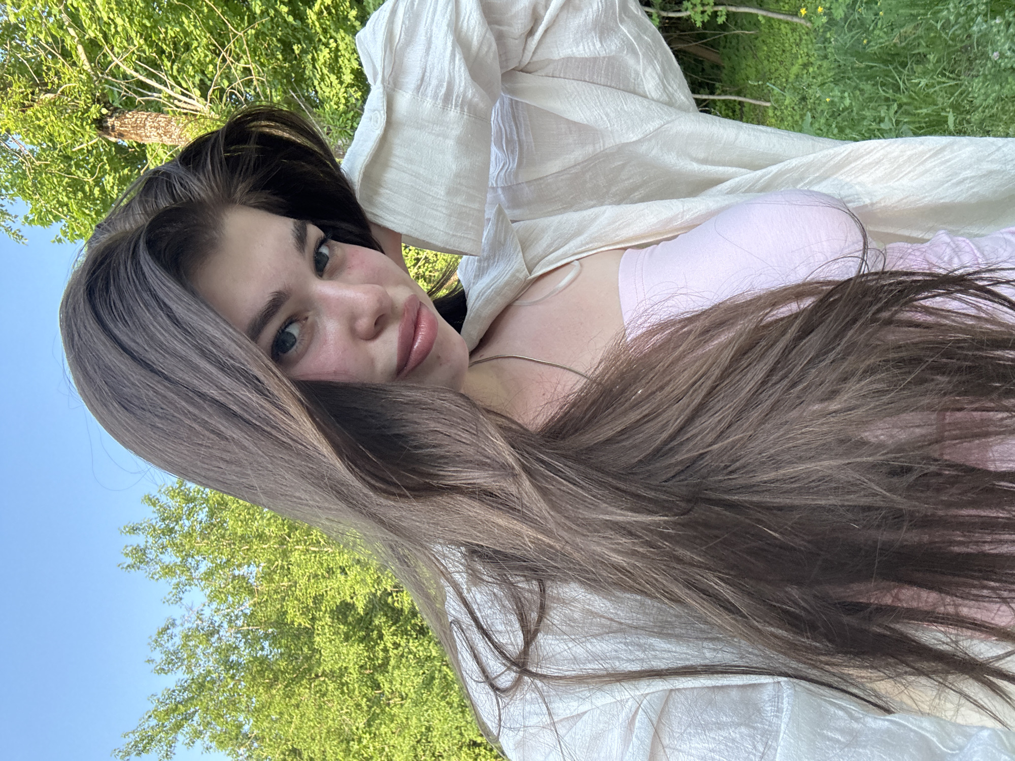 A young woman with long brown hair is positioned in the foreground of the image. She is wearing a pink top and a white textured shirt. The background features greenery including grass, trees and blue sky, possibly indicating a natural setting such as a park.
It is not possible to confirm the image was taken specifically in the city of Посёлок комбината стройматериалов-2, Russia. The background lacks identifying features to determine the specific location.