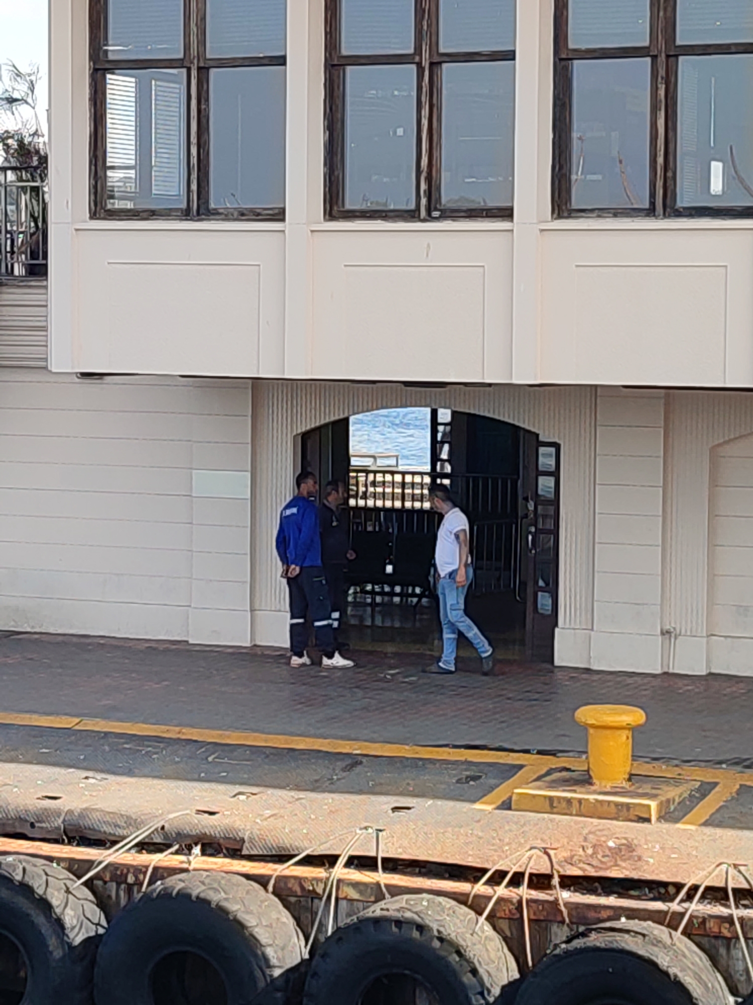 In Kadıköy, Turkey, a scene unfolds outside a building with large windows. Three men are present. Two men, wearing blue and dark-colored work uniforms respectively, stand near an arched entrance, appearing to converse. A third man, in a white t-shirt and blue jeans, walks away from the entrance. The ground is paved, with yellow lines marking a path, and a yellow bollard is visible. In the foreground, large tires are stacked on a dock-like structure. The building's facade is cream-colored, with decorative vertical fluting.