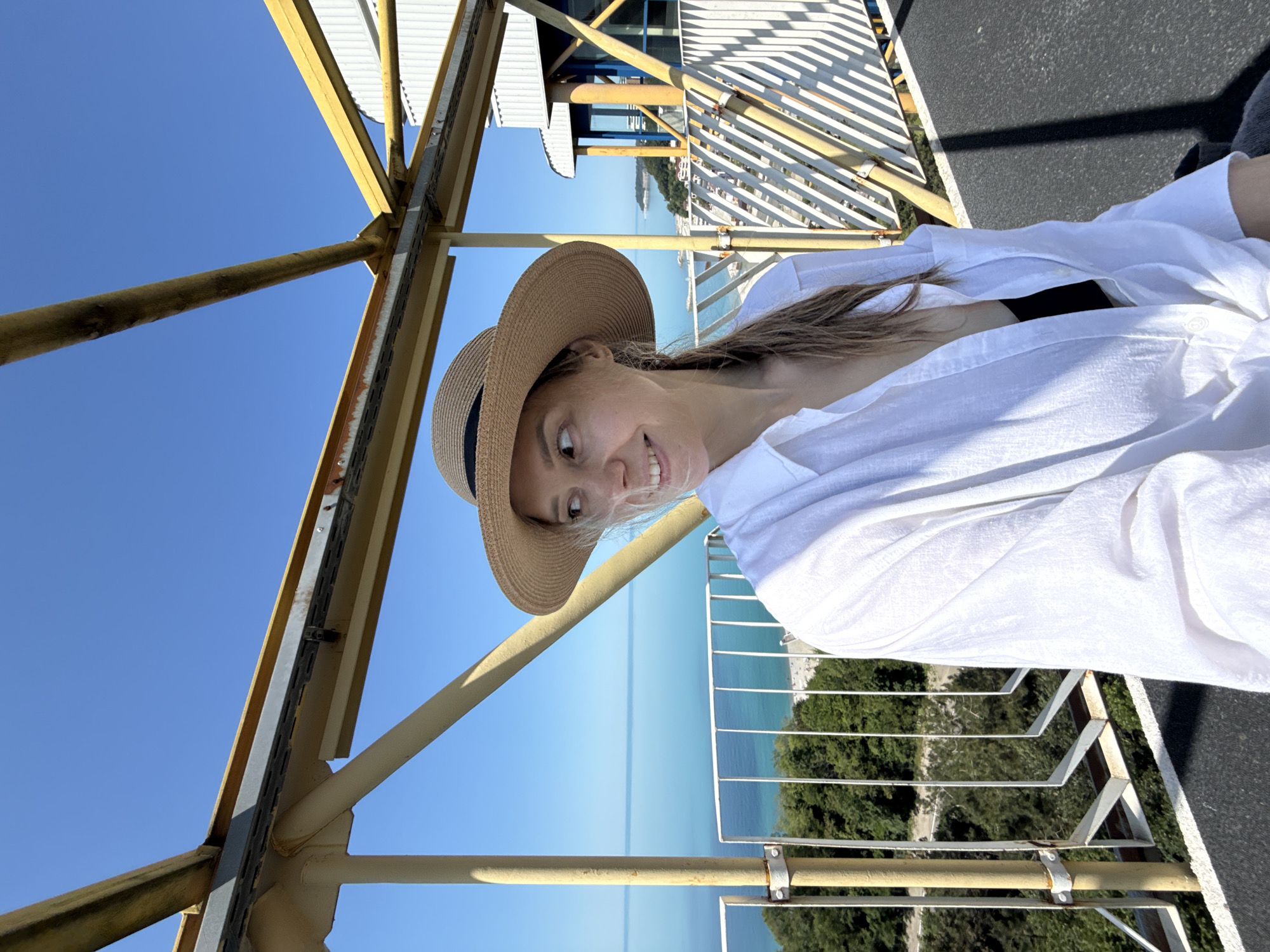 An adult female is centrally positioned on an elevated outdoor platform under bright sunlight in Nebug, Russia. She wears a wide-brimmed straw hat with a dark band, an unbuttoned white long-sleeved shirt revealing a black top, and her light brown hair is tied back. Her head is turned to her left (viewer's right), displaying a slight smile. The platform is constructed with yellow metal beams, vertical supports, and a white metal railing. The surface beneath her is dark and textured. In the background, a clear blue sky extends over the Black Sea, which transitions from a turquoise hue near the visible pebbled coastline to a deeper blue further out. Green trees and a winding road are visible below the platform, descending towards the beach. To the right, sections of buildings with white siding and blue windows are partially visible beyond the platform's structure, indicative of coastal architecture.