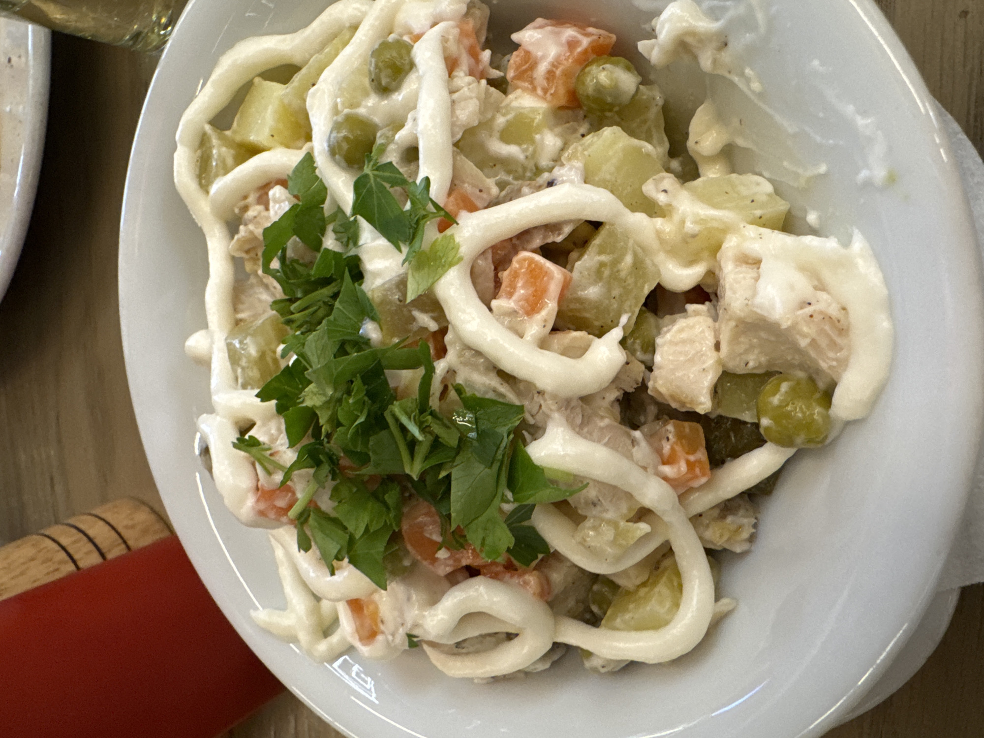 A top-down, close-up shot shows a white plate filled with Olivier salad. The salad consists of diced potatoes, carrots, green peas, what appears to be chicken or ham, and is dressed with mayonnaise. A pile of chopped fresh green herbs, possibly parsley, sits on top of the salad. Parts of the plate are smeared with the salad dressing. In the lower-left corner, there is a glimpse of what appears to be a red object with a dark-stained wooden handle or accent. A second plate is shown on the side, but is out of focus. The image, given the dish, may be located in Yerevan, Armenia, where Olivier salad is a common dish, but there are no specific identifiers to confirm the location. The image lacks any people, actions, or interactions.