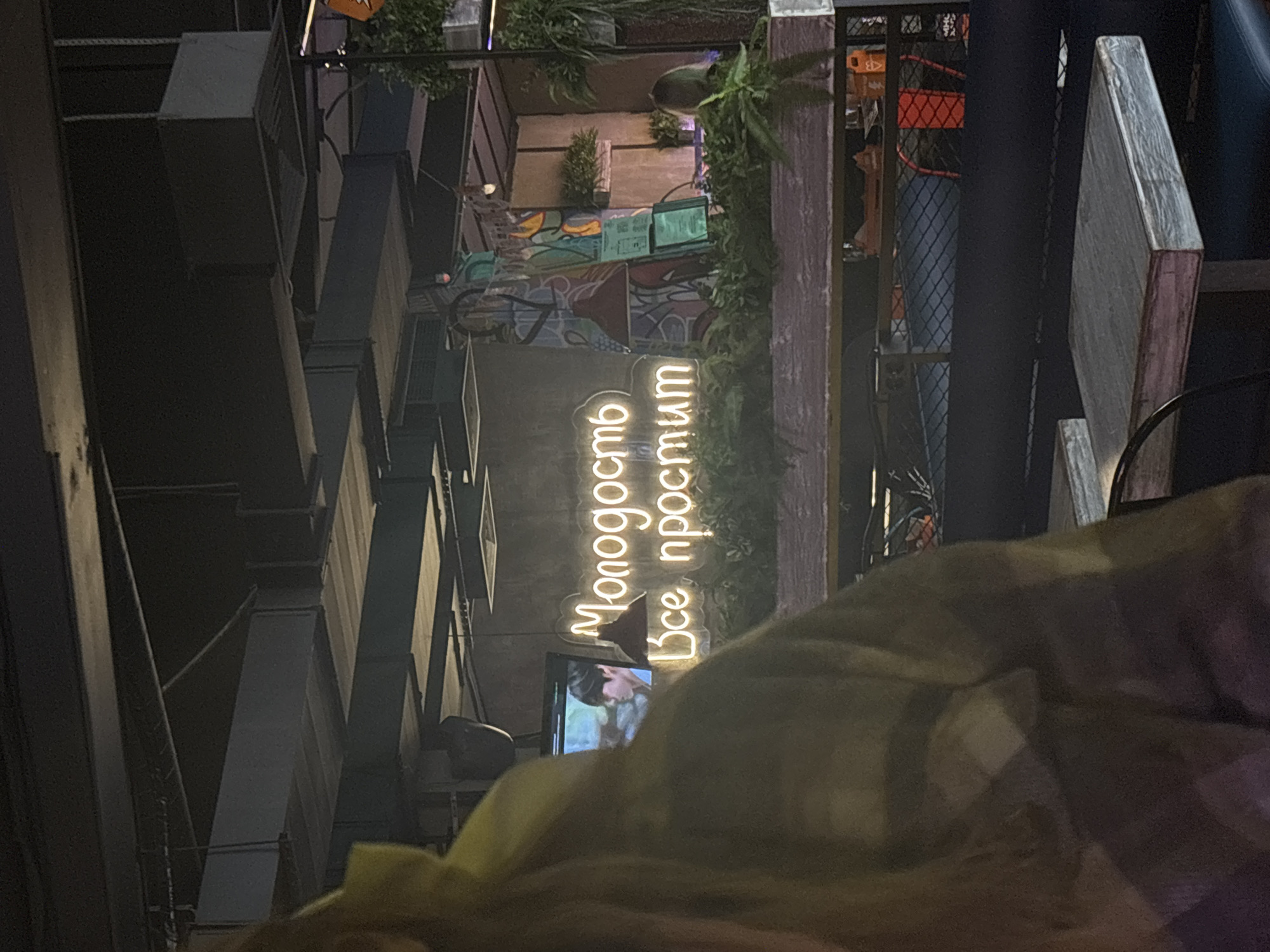 The image, likely taken in Saint Petersburg, Russia, shows an exterior shot of a building with architectural details including metal beams, ledges, and planters. A neon sign in Russian, reading "Молодость Всё простит" (Youth Forgives Everything), is prominently displayed. A television screen is also visible below the sign. The view is partially obstructed by a blurry object or person in the foreground. The lighting suggests it is either nighttime or a dimly lit environment.