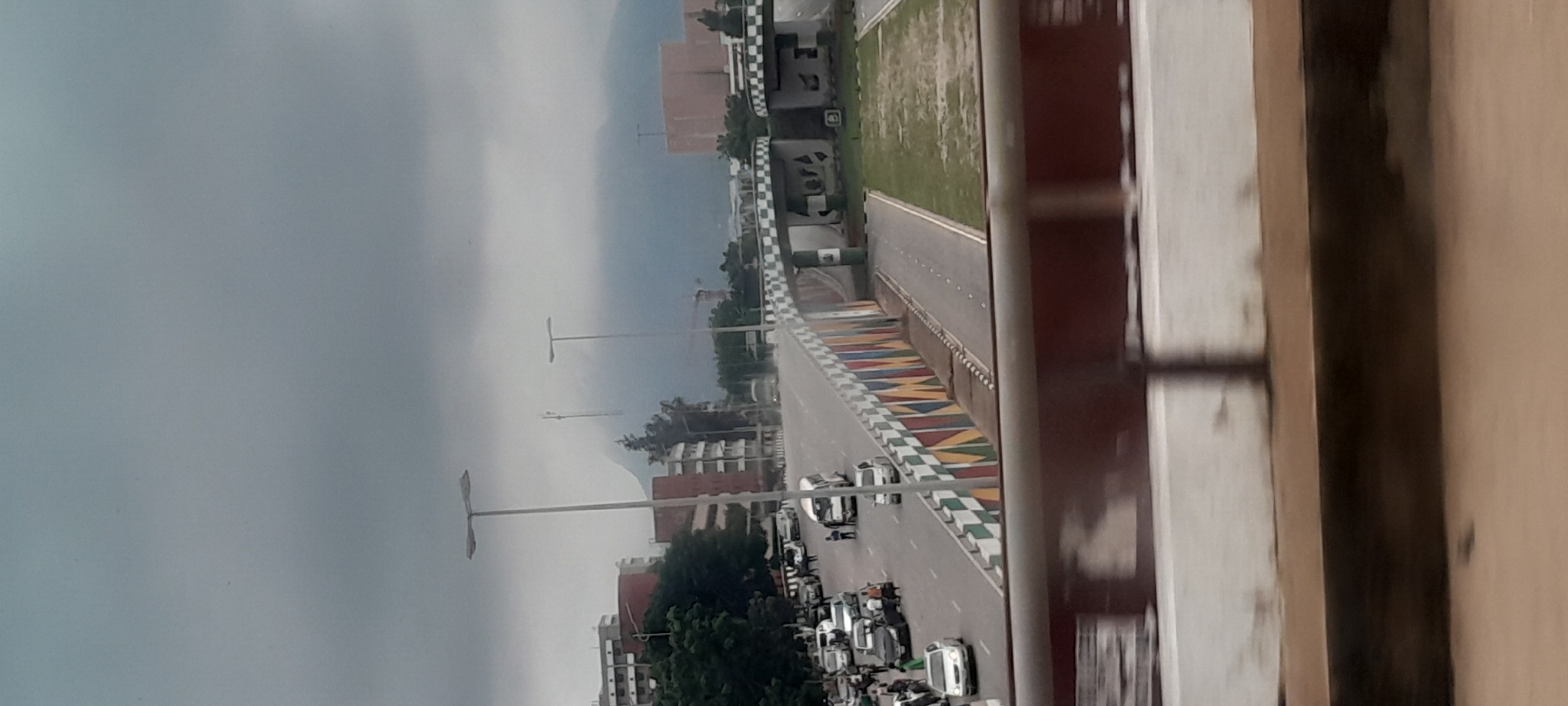 An overcast, cloudy day frames a view of a multi-lane road in Abuja, Nigeria, with buildings and a mountain range in the background. The road appears to be elevated, with a decorative bridge railing adorned with colorful geometric patterns and a checkered design. Several vehicles, including cars and a white van, are visible on the road, with some people standing near them. Tall streetlights are positioned along the road. The architecture of the buildings suggests an urban environment, and the presence of communication towers and a construction crane hints at ongoing development. The foreground shows a blurred railing, indicating the photo was taken from a moving vehicle. The overall atmosphere is subdued, possibly due to the cloudy weather.