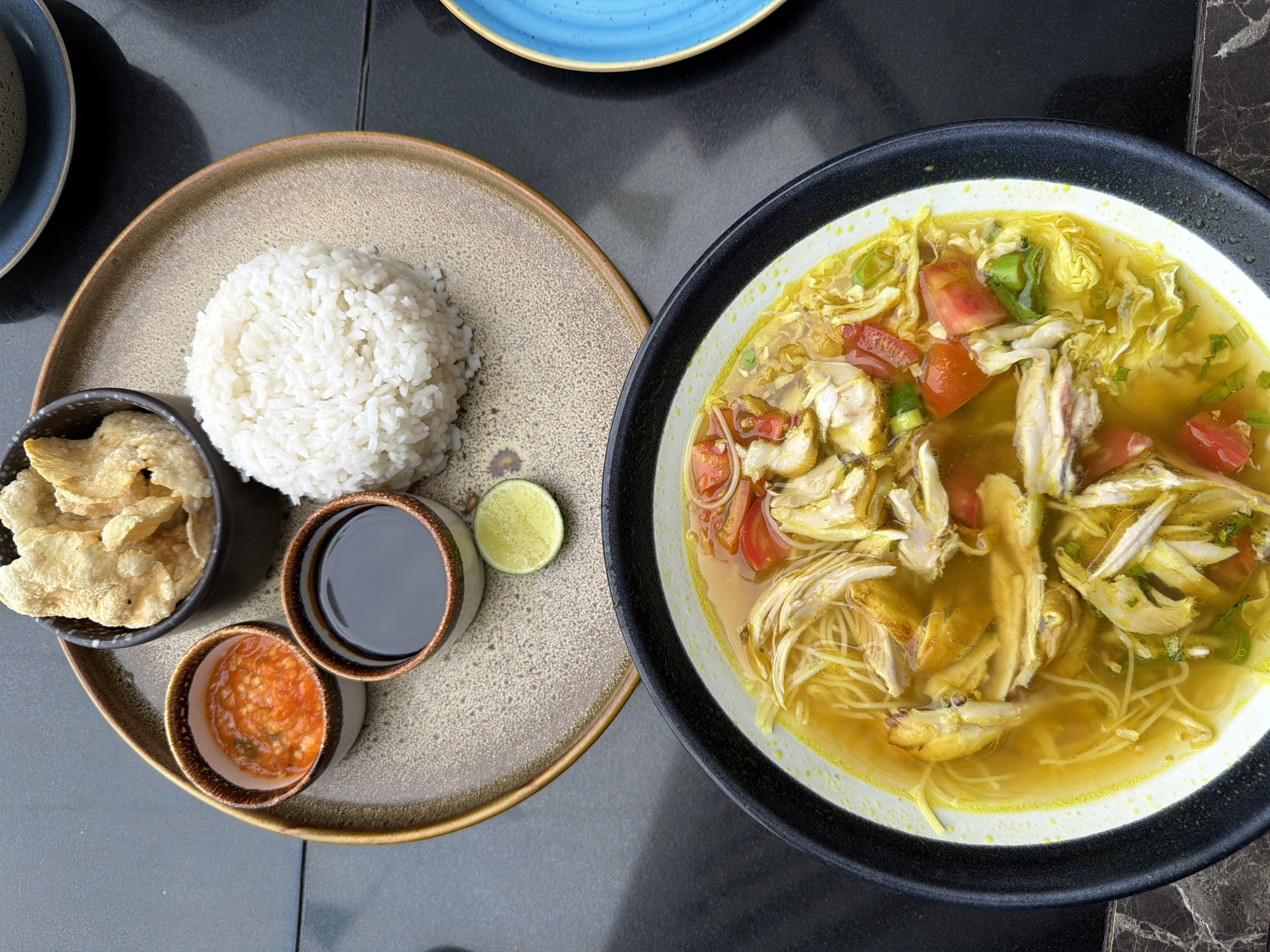 The image shows a meal of Soto Ayam, a traditional Indonesian chicken soup, served with rice and condiments. The soup is in a black-rimmed bowl and appears to contain shredded chicken, noodles, and diced tomatoes in a yellow broth. On a separate plate, there is a mound of white rice, a small bowl of what appears to be shrimp crackers, a small bowl of soy sauce, a small bowl of red chili sauce, and a lime wedge. The picture is taken from an overhead perspective, likely at a restaurant in the city of South Jakarta, Indonesia. The table appears to be dark-colored, possibly stone or a similar material.