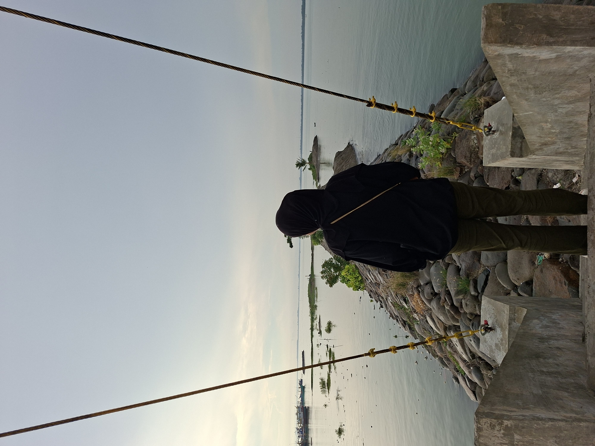 In Lhokseumawe, Indonesia, a person wearing a black hijab and a black hoodie stands on a concrete staircase, facing away from the camera. They are looking out at a body of water that extends to the horizon. The water is calm and reflects the overcast sky. A narrow, rocky path, possibly a breakwater or jetty, extends into the water. Small islands with sparse vegetation are visible in the distance. Several boats are anchored or moving in the water to the left of the frame. Two thick ropes, attached to concrete structures, extend diagonally upwards from the foreground on either side of the person, creating a visual frame. The overall scene depicts a serene, coastal environment under a diffused sky.