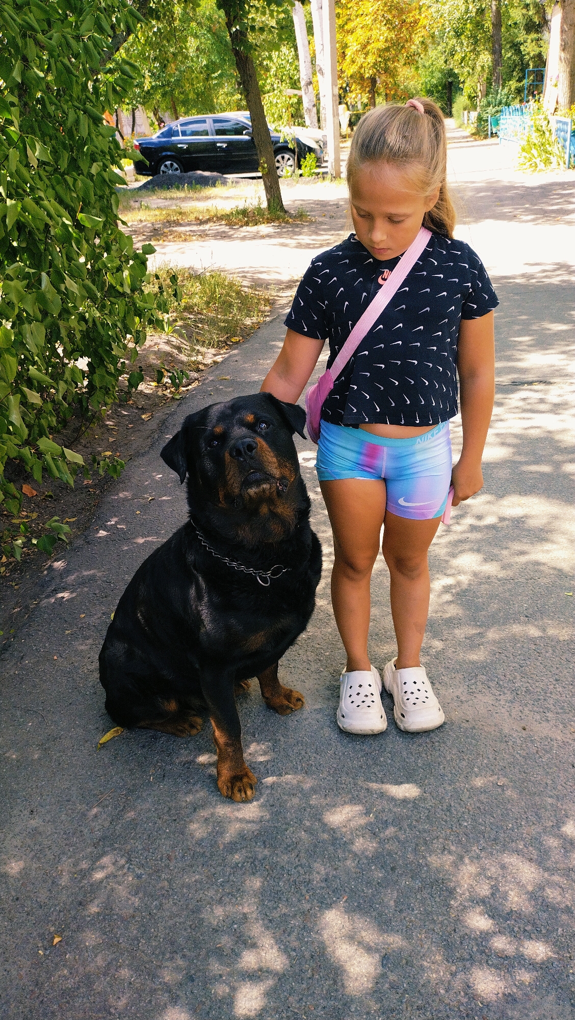 A young girl with blonde hair in a ponytail, wearing a dark t-shirt with a white pattern and colorful athletic shorts, stands on a paved path. She has her right hand resting on the head of a Rottweiler dog sitting beside her. The dog is wearing a silver chain collar. The scene appears to be outdoors in a park-like setting, with trees and foliage on the left and in the background. A black car is visible in the distance behind the trees. The lighting suggests it is daytime. This image was taken in Horishni Plavni, Ukraine.