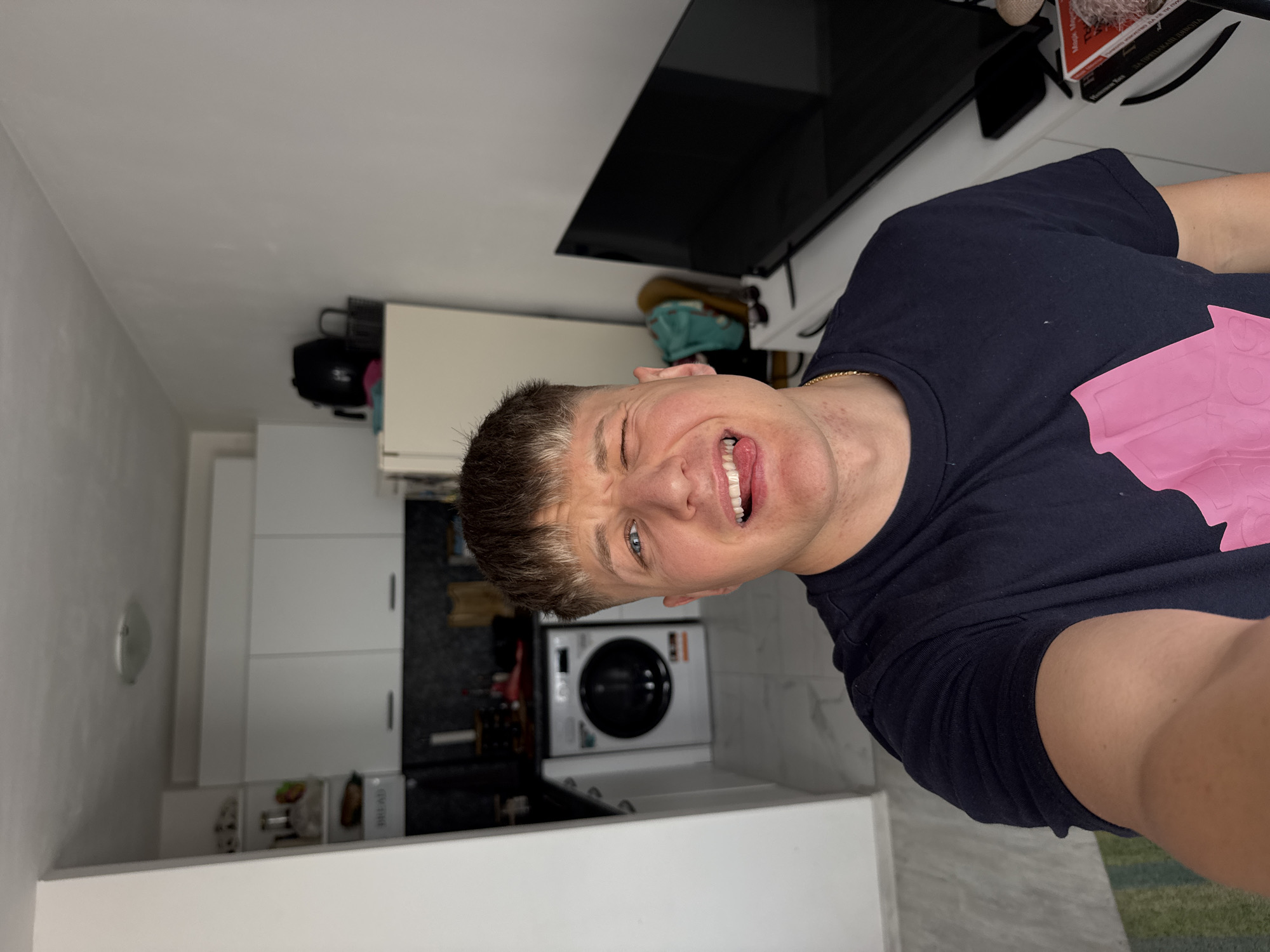 A man stands in a small kitchen in Plovdiv, Bulgaria. He is Caucasian, fair-skinned, with short brown hair, blue eyes, and a gold chain. He is wearing a dark t-shirt with a pink design. He is sticking his tongue out and squinting one eye.
The kitchen features white cabinets, a washing machine, and a black tiled backsplash. A television is mounted on the wall. Items are stored on top of the cabinets, including a black bag and various containers. The room is well-lit.
