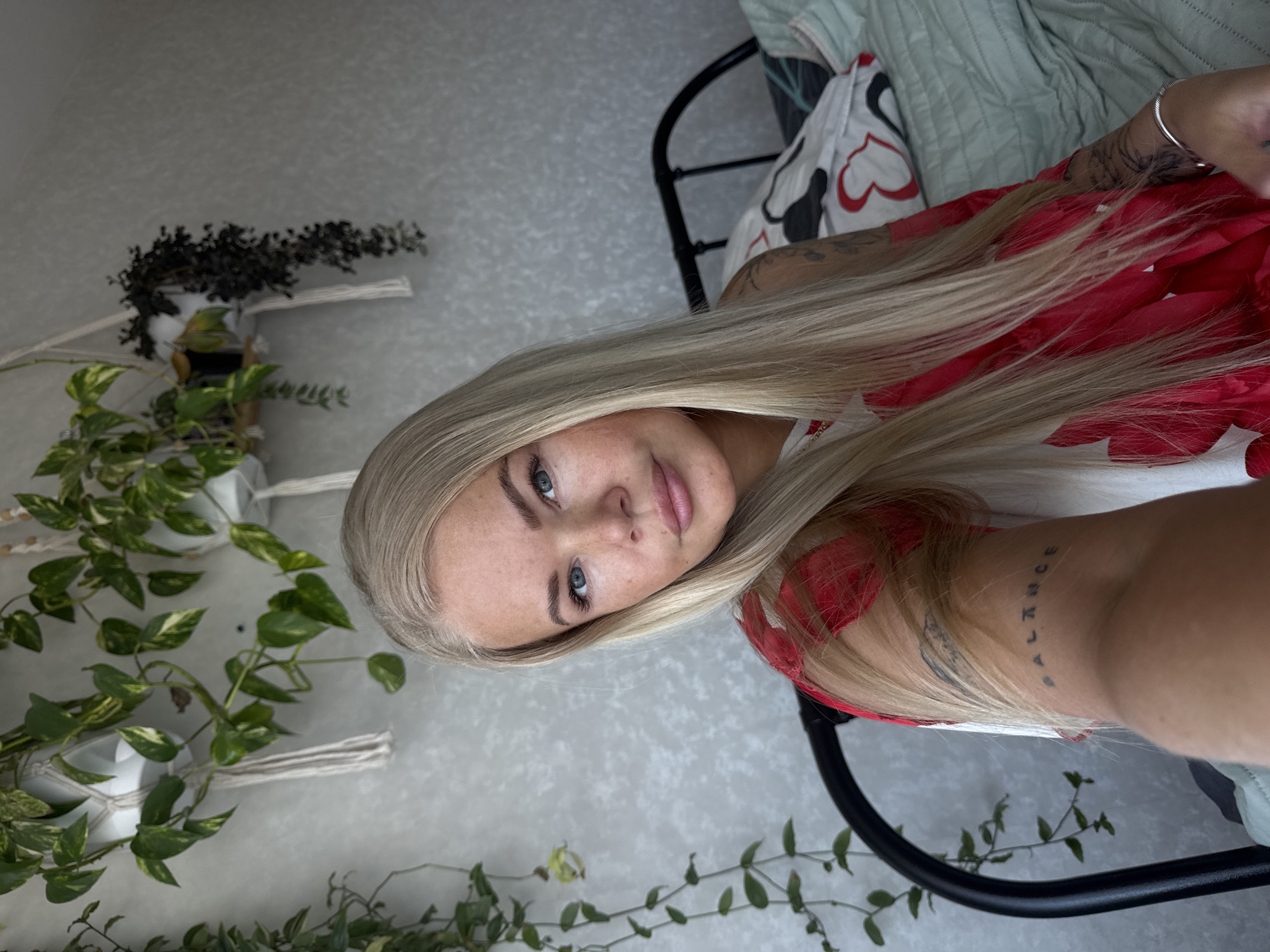 A fair-skinned woman with long, straight blonde hair is centered in a close-up shot, seemingly indoors. She has blue eyes and is wearing a red floral print top. Her left arm is visible and shows tattoos. Behind her, a black metal bed frame is partially visible, with bedding that includes a pillow or blanket with a white and red pattern. To her left are hanging plants in macrame planters, and other vines are also present in the background. The background wall is a textured light gray.