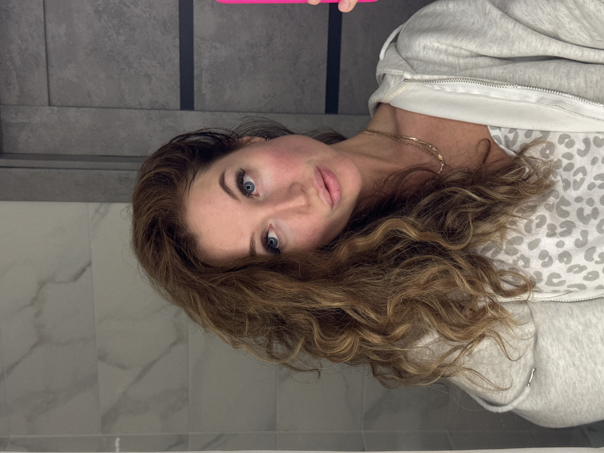 This close-up selfie features a woman with light brown, wavy hair and striking blue eyes, captured in an indoor setting. She has fair skin, highlighted by a rosy blush on her cheeks, and full, lightly glossed lips. Her left eye displays prominent eyelash extensions. She is dressed in a light grey zippered hooded sweatshirt, with a white and grey leopard print top visible underneath, and a thin gold chain necklace adorns her neck. A bright pink object, likely a smartphone, is partially visible at the top of the frame, held by her hand as she takes the photo. The woman looks directly at the camera with a calm, neutral expression.
The background consists of tiled walls. The upper section features dark grey, rectangular tiles, while the lower section showcases lighter grey tiles with a subtle marble pattern, separated by a dark line. A white horizontal line is visible at the very bottom of the frame, suggesting a countertop or bathtub edge. The image is oriented sideways, with the woman's head tilted. The scene takes place indoors, likely within a bathroom or a similar private space. No specific time of day or weather conditions are discernible. The location of the photograph is attributed to Chaikovsky, Russia. There is no visible text in the image.