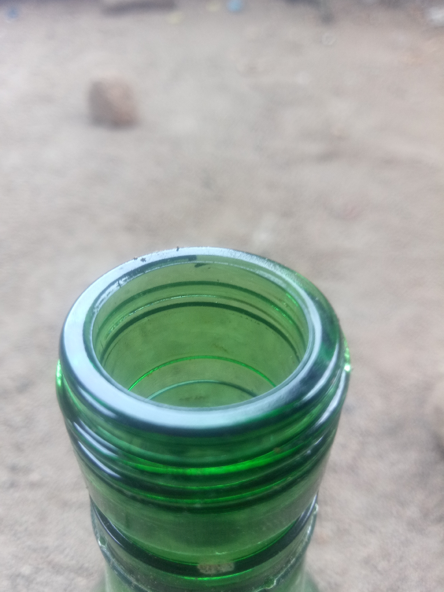 A close-up, high-angle, vertical shot shows the opening of a green glass bottle. The bottle has a thick rim with several concentric ridges around the neck. The interior of the bottle is visible and appears to be empty and clean. The background is out of focus and appears to be a sandy or gravelly surface, possibly outdoors. A blurred, light-colored object is visible in the upper left portion of the background. The lighting is natural and the overall scene suggests an outdoor setting in a non-urban environment.
