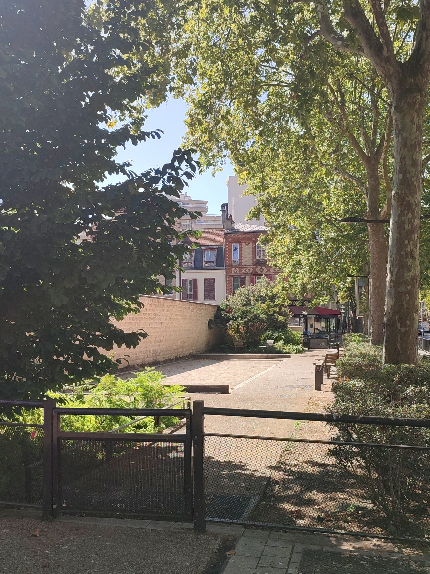 The image depicts a street scene in Boulogne-Billancourt, France. In the foreground, a dark metal fence with mesh panels separates a gravel path from a paved walkway. Beyond the fence, lush green foliage is visible. Further back, a light-colored brick wall runs horizontally across the mid-ground. A paved area with what appears to be a bocce ball court is situated behind the wall. Several wooden benches are placed along the side of this paved area.
In the background, buildings with red-tiled roofs and red-shuttered windows are visible. One building has a red awning extending over a street-level establishment labeled "BAR BRASSERIE". Tall trees with dense green foliage line the right side of the scene, casting dappled shadows on the ground. The sky above is clear and blue. The overall impression is a pleasant, sunny day in an urban environment with green spaces and residential buildings.