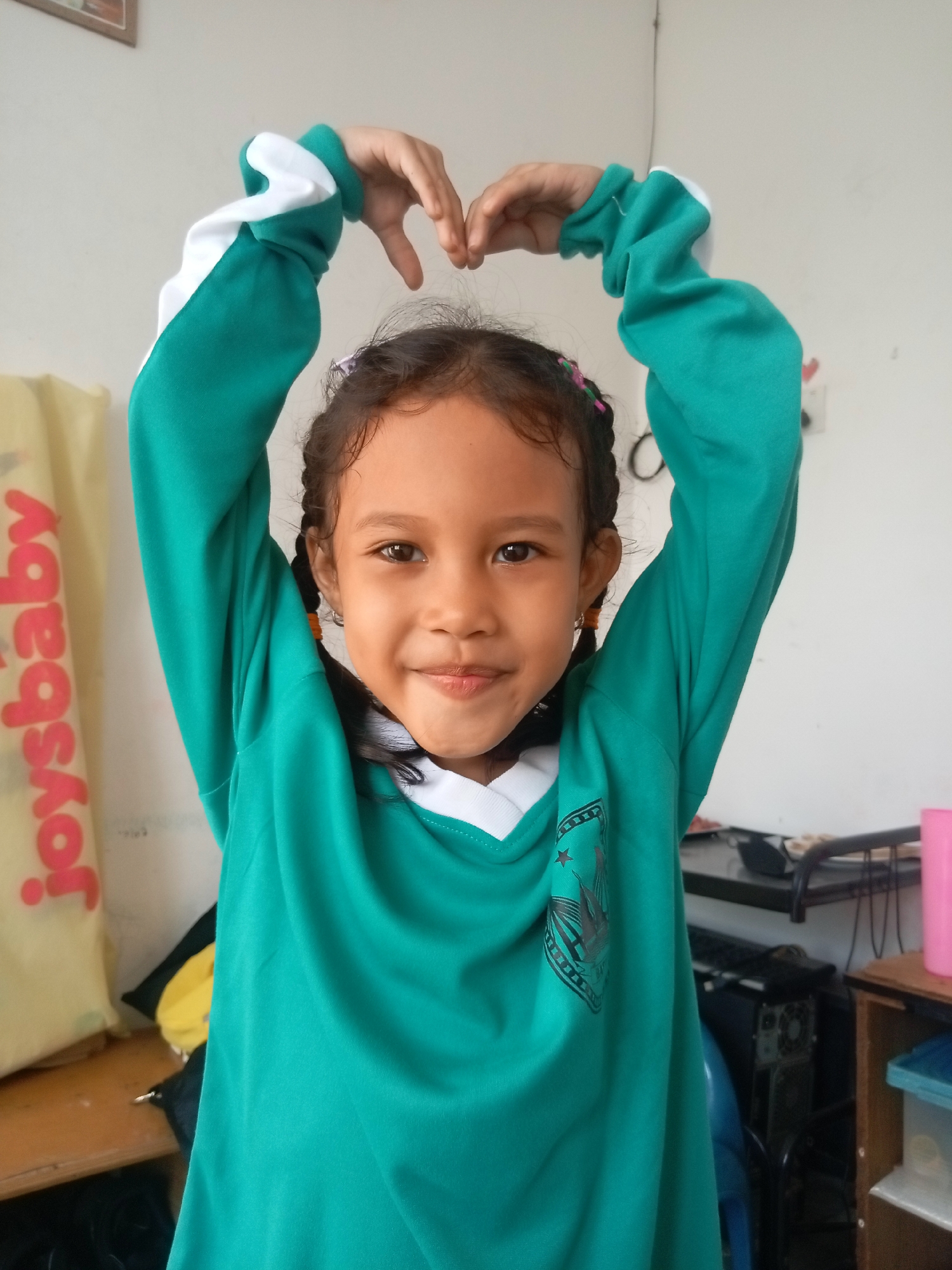 This media file is a portrait of a young girl, likely of Indonesian descent given the Batam location context. She has dark hair styled in two braids, with a few colorful hair accessories visible. The girl is wearing a long-sleeved, teal and white v-neck shirt. She is smiling warmly at the camera, her eyes bright and engaging. Her hands are raised above her head, with her fingers touching to form a heart shape. The background is a plain, light-colored wall, with some objects visible to the left and right. To the left, a yellow bag with red text that appears to read "joysbaby" is partially visible. To the right, there are shelves with various items, including what looks like a computer tower, a pink cup, and some containers. The lighting suggests it is daytime, possibly indoors. The overall mood is cheerful and positive, captured by the girl's bright smile and the heart gesture.
