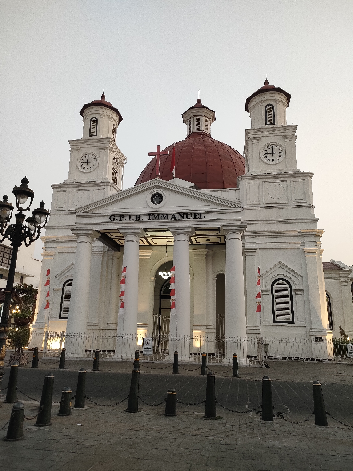 The image shows a frontal view of GPIB Immanuel, a white church with a red dome and two clock towers, located in Semarang, Indonesia. The church features classical architecture with large white columns and decorative elements. Red and white flags are draped on the facade. In the foreground, there is a paved street with bollards and chains, and a lamppost to the left. The sky is overcast and the light suggests it is either early morning or late afternoon. There are no people visible in the image.