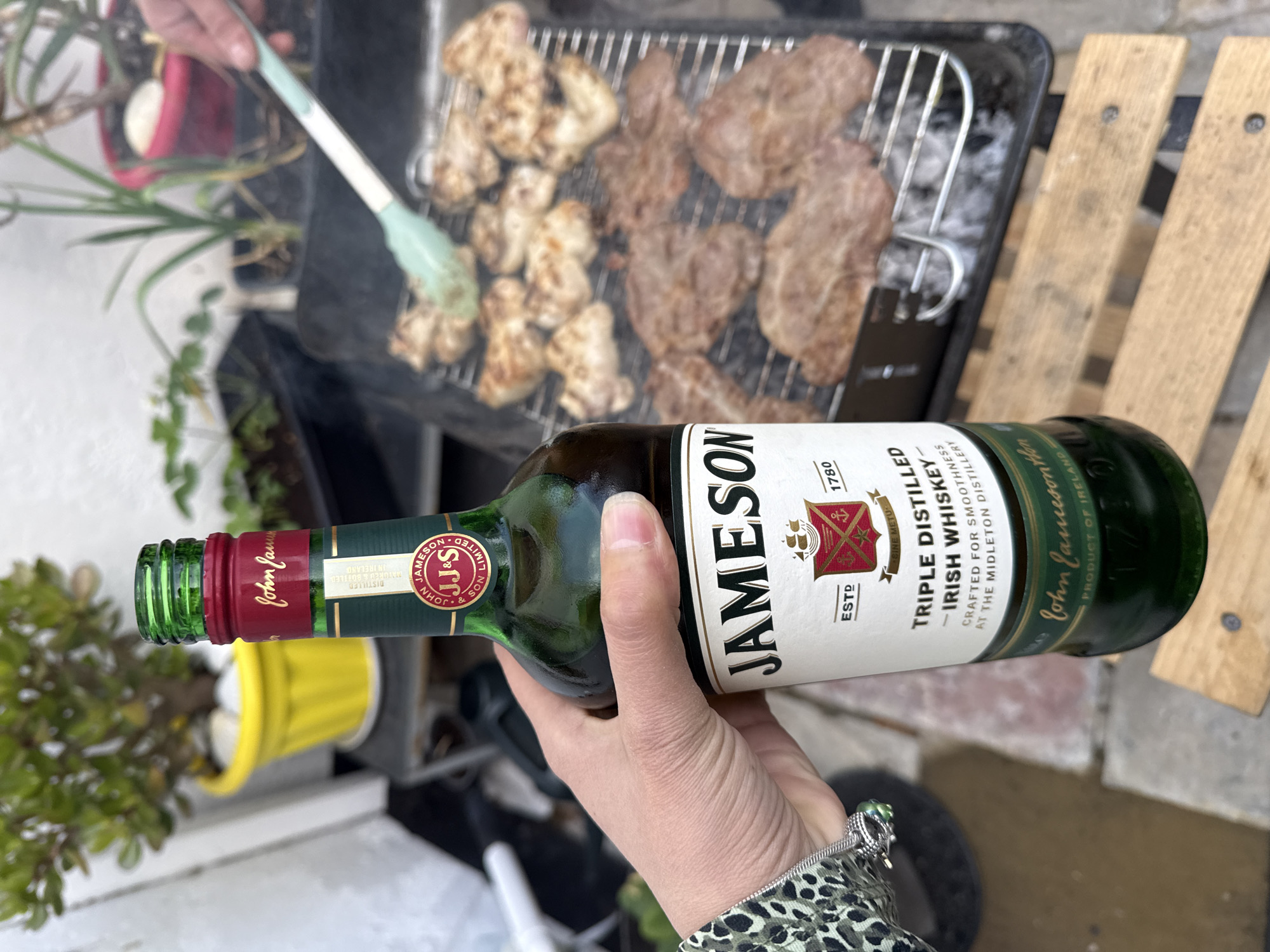 The scene is an outdoor cooking area. A hand holds a bottle of Jameson Irish Whiskey. In the background, a grill displays cooked meat and chicken. The surroundings include plants in pots, and wooden slats. The specific location in Great Yarmouth, United Kingdom, cannot be determined from the visual information.