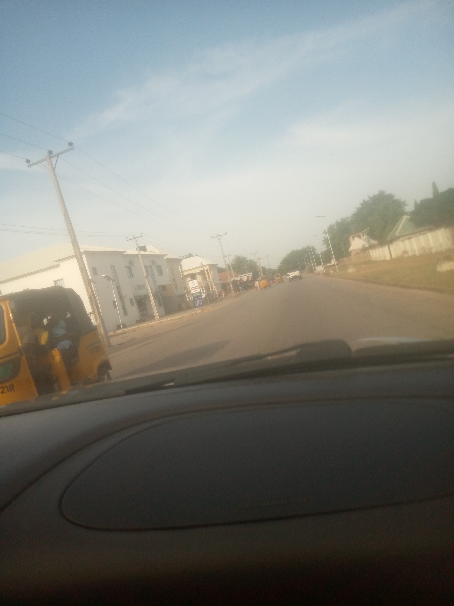 The image captures a scene in Jalingo, Nigeria, likely during the day with clear skies. The perspective is from inside a vehicle, looking forward through the windshield at a road.
In the foreground, a dark dashboard is visible, suggesting the vantage point of a driver or passenger. To the left, a yellow motorized tricycle, commonly known as a "keke," is parked or moving slowly. A person is visible inside the keke, appearing to be a passenger or driver. The keke has some visible text, "21R," on its side.
The road stretches into the distance, lined with buildings on both sides. On the left, there are modern, white-painted buildings, some appearing to be multi-story. A prominent utility pole with electrical wires stands near the keke. Further down the road, more buildings are visible, along with other vehicles, including what appears to be a white pickup truck and possibly other tricycles.
On the right side of the road, a low wall with a fence runs along the edge of the property. Trees and greenery are visible behind the wall, and more buildings can be seen in the background, some with light-colored walls and tiled roofs. The overall atmosphere suggests a typical day on a street in this Nigerian city, with everyday vehicles and architecture. The lighting indicates it is daytime, possibly morning or afternoon, with bright, diffused sunlight. No strong emotional cues are evident in the image.