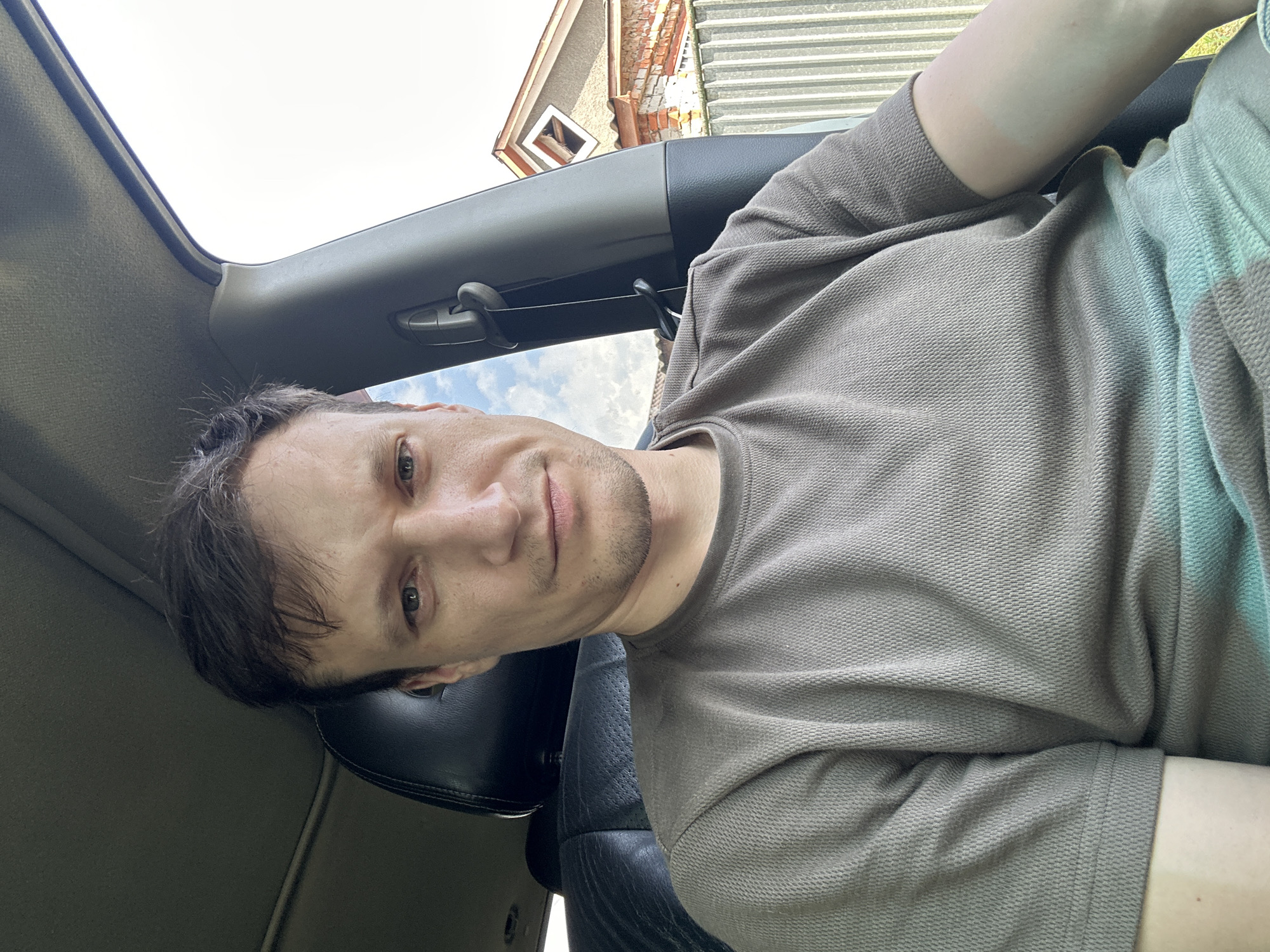 A man with short brown hair and a neutral expression is seated in the driver's seat of a vehicle, looking directly at the camera. He is wearing a brown t-shirt. The interior of the car, including a black leather seat and part of the dashboard, is visible. Outside the car, a pale blue sky with some clouds can be seen, along with a fence and a building with a red-tiled roof. The image appears to be taken outdoors, possibly on a street in the city of Makiv, Ukraine.