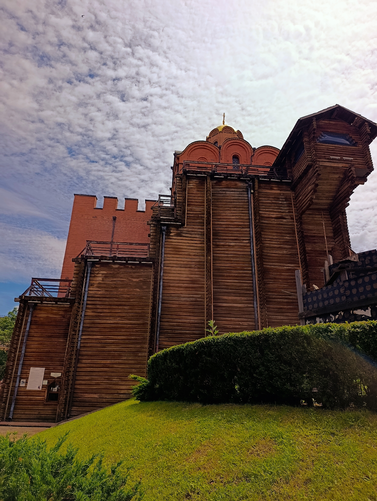 The image captures the Golden Gate in Kyiv, Ukraine. The structure is predominantly made of wood with brick elements and features a golden dome. It is surrounded by lush green grass and manicured hedges. The sky above is filled with scattered white clouds, and a few birds are visible in flight. The perspective is from a low angle, looking up at the gate.