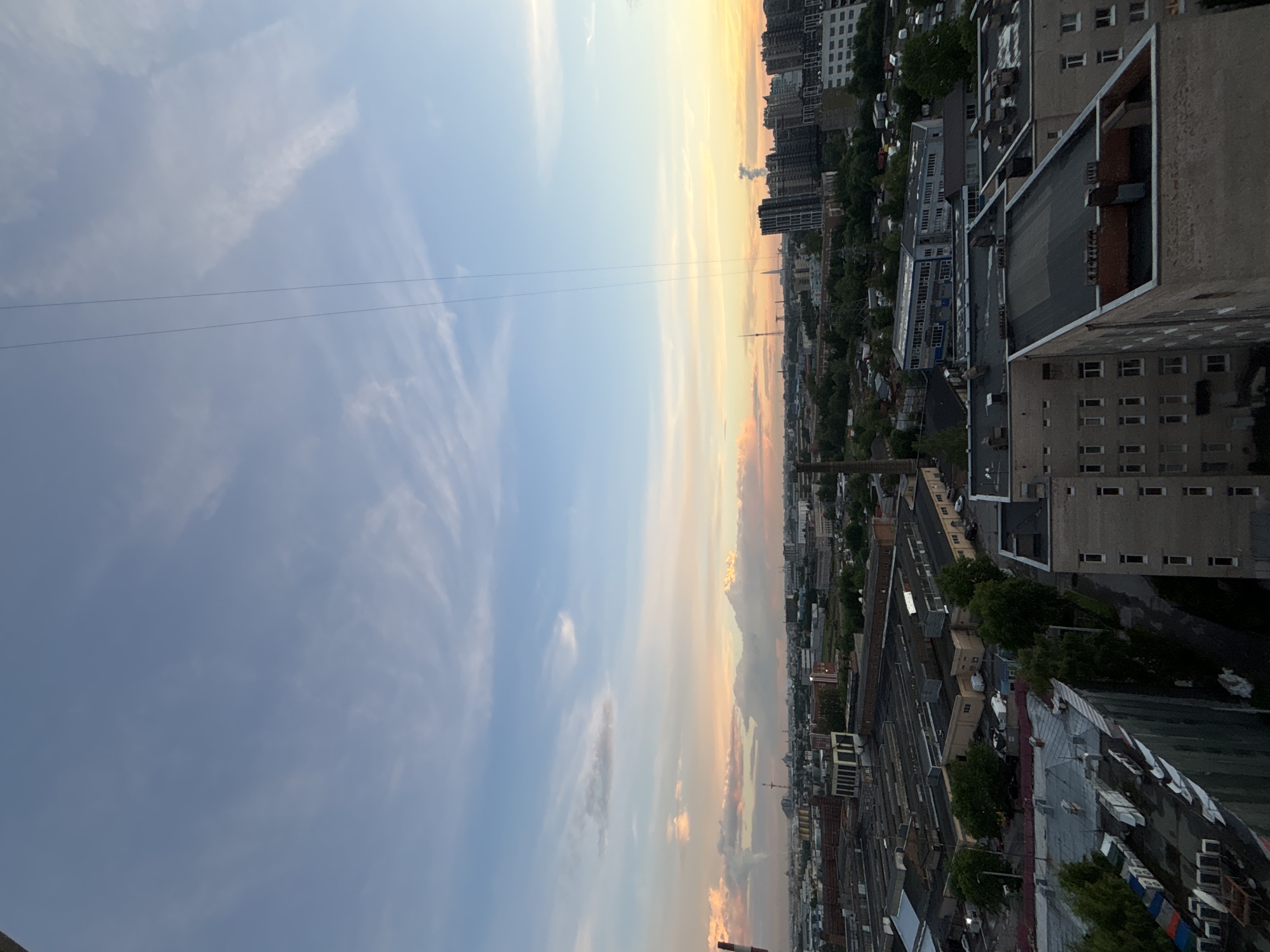 The image depicts an aerial view of an urban industrial landscape during a sunset. The sky is a gradient of blues, purples, and oranges, with scattered wispy clouds. Two thin, dark wires extend vertically down from the top of the frame, likely power lines or antenna wires.
Below the sky, a city sprawls with a mix of industrial buildings, warehouses, and residential high-rises. The industrial section in the foreground features long, low-lying factory-like buildings with corrugated metal roofs and brick chimneys. Some of these buildings appear to be older, with visible signs of wear. Further into the distance, taller, modern apartment buildings with dark facades are visible. Green trees are interspersed throughout the cityscape, particularly around the residential areas. The overall impression is of a dense, developed urban environment, possibly on the outskirts of a major city, given the presence of both industrial and residential structures. The lighting suggests it is late evening.