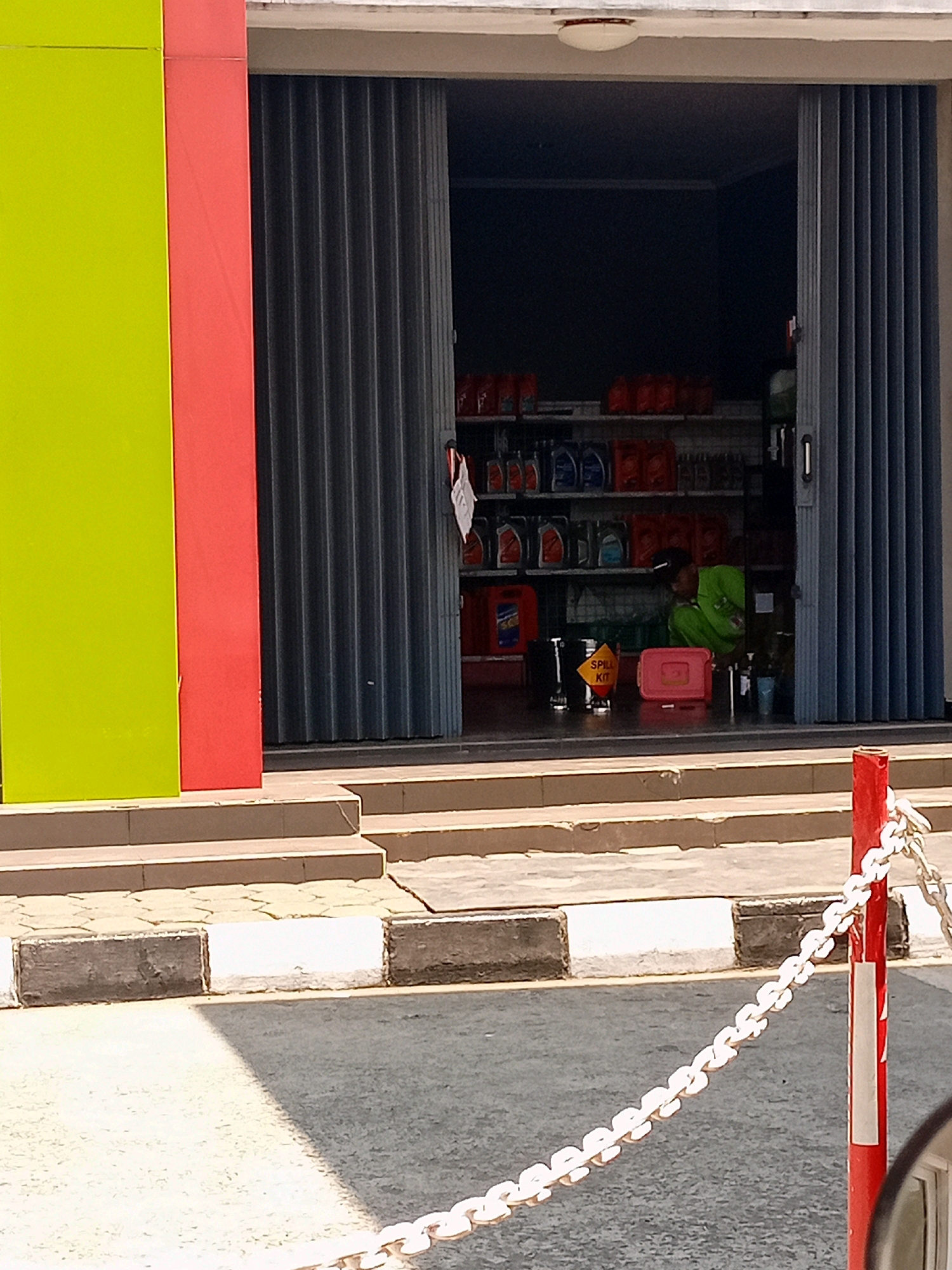 A person wearing a green shirt and black pants is squatting inside a store that sells various automotive fluids. The store has shelves stocked with numerous bottles and containers of oil and other liquids. A red plastic container and a yellow sign with "SPILL KIT" written on it are visible on the floor in front of the person. The store entrance is a large, gray, collapsible metal gate. Outside the store, there are steps leading up to the entrance, and a street with a white and gray curb. A red pole with a white chain attached is also visible in the foreground, to the right of the image. The store has a bright green and red exterior. The scene appears to be located in a commercial area, possibly a small town or city.