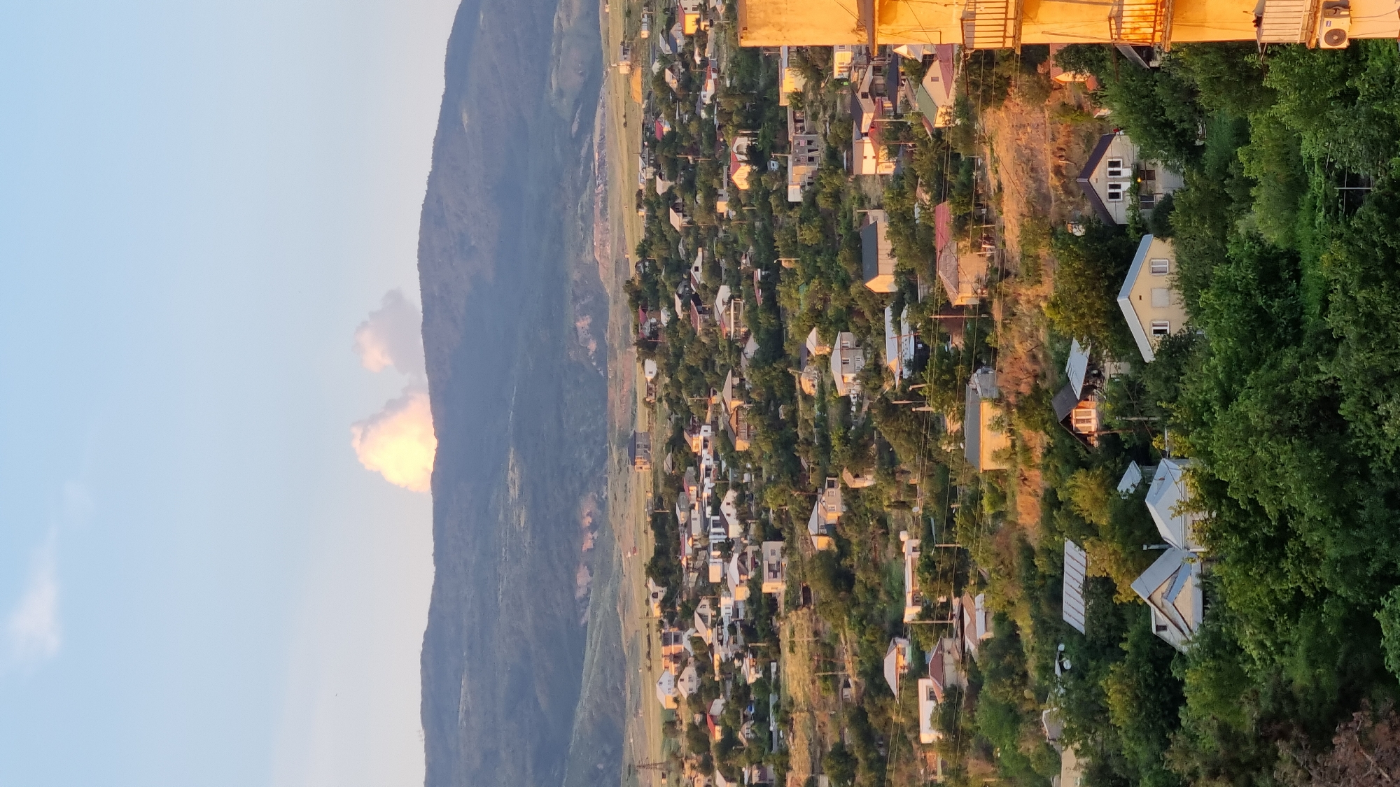 The image captures a panoramic view of a hillside residential area in Tbilisi, Georgia, bathed in the warm glow of what appears to be late afternoon or early evening sunlight. Dominating the upper portion of the frame are rolling, forested hills and mountains, with a dramatic, bright cloud formation, seemingly lit from within, positioned directly above the highest peak. The sky is a clear, pale blue, with a single smaller cloud visible to the left of the larger formation.
The midground is densely populated with numerous houses, varying in size and architectural style, nestled amongst lush green trees and vegetation. The houses are primarily single-family dwellings, many featuring light-colored exteriors and pitched roofs. Some appear to be older, more established structures, while others suggest ongoing construction or recent renovations. Roads and pathways wind through the settlement, and utility poles with visible wires crisscross the scene, indicating the presence of infrastructure.
The foreground showcases more dense foliage, with the tops of trees creating a verdant border across the bottom of the image. On the far right of the foreground, a portion of a building, possibly an apartment block, is visible. This building is a light yellow-tan color and has balconies with railings, suggesting a different type of urban development compared to the houses on the hill. An air conditioning unit is visible on the exterior wall of this building.
The overall impression is of a peaceful, established residential community situated within a natural, hilly landscape, characteristic of many areas in Tbilisi. There are no people or overt human activity depicted in the image. The focus is on the built environment and its integration with the natural scenery, highlighted by the atmospheric lighting.