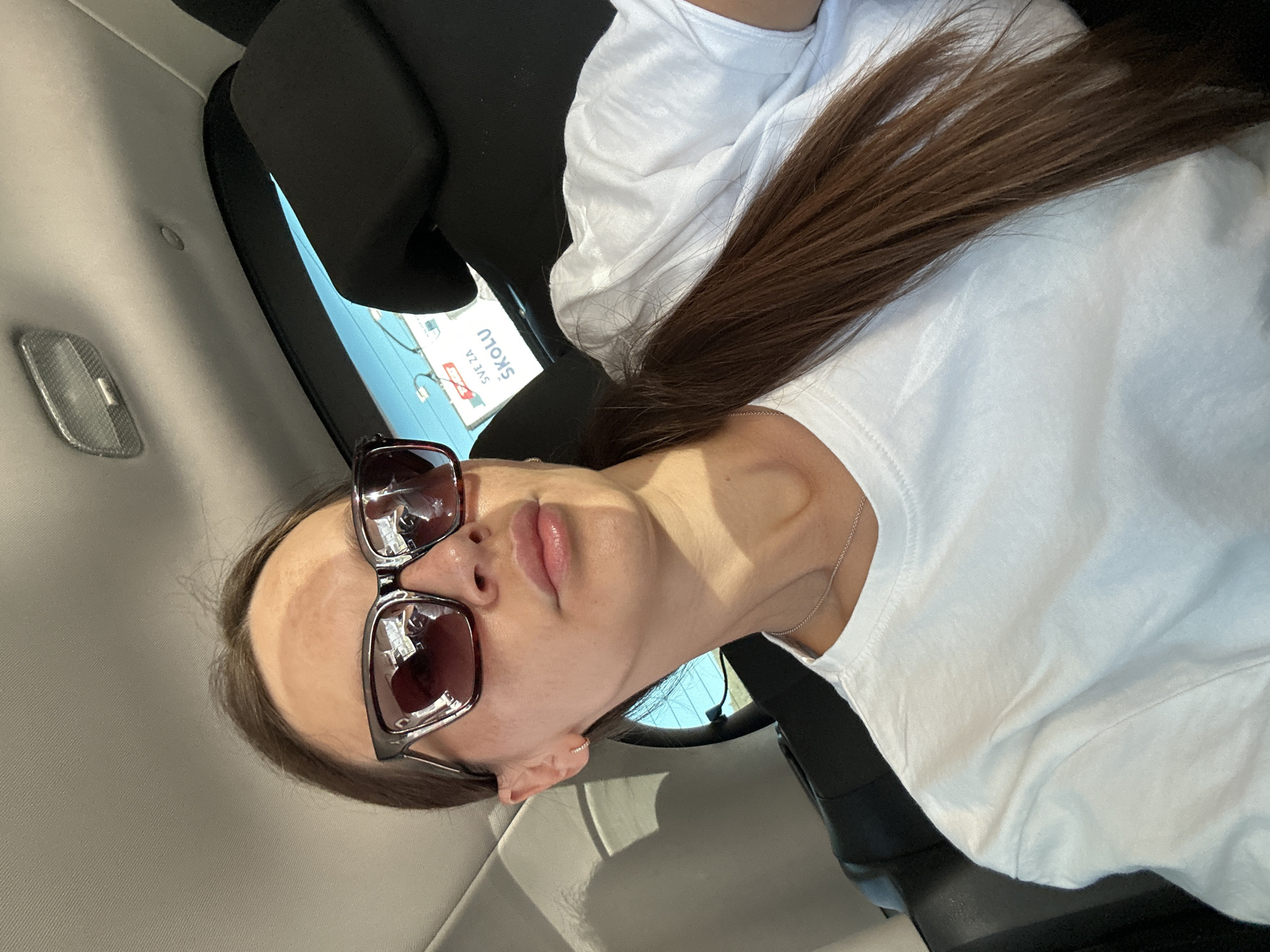 A young woman with long brown hair pulled into a ponytail is taking a selfie in a car. She is wearing sunglasses and a white t-shirt. The interior of the car is visible, including the headrests and the ceiling. Outside the car window, a blue sky and some trees can be seen. A sign is also visible through the window with text that appears to be in a Slavic language, possibly advertising school supplies. Based on the visible text, the location is likely Budva, Montenegro, as "Sve za školu" translates to "Everything for school" and is a common slogan for school supply stores.