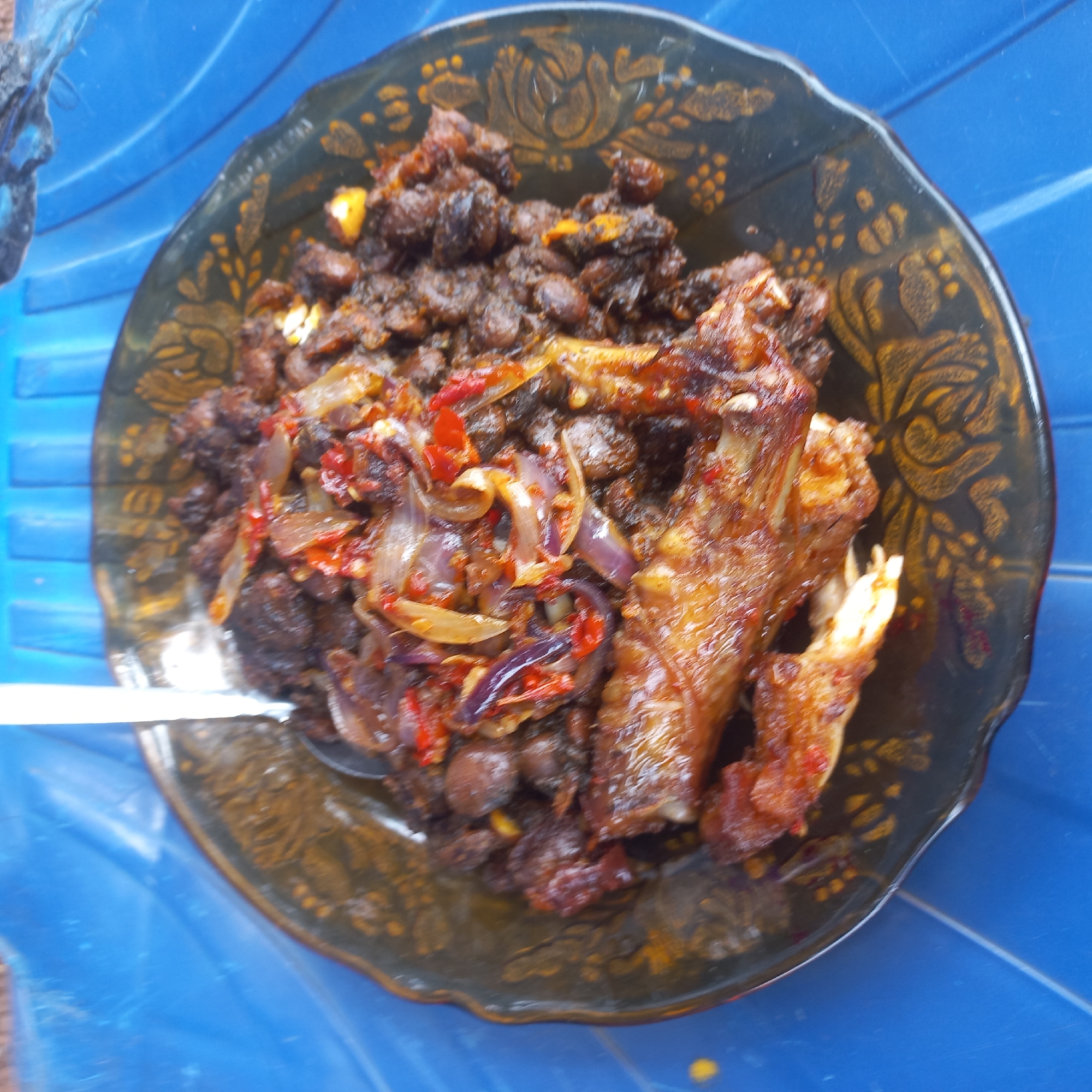 A plate of food containing what appears to be fried meat, onions, and chili peppers is presented on a blue surface. The plate has a decorative rim with an intricate, embossed pattern in gold and black. A metal utensil, possibly a fork or spoon, is visible on the left side of the plate, partially submerged in the food. The food itself is a dark brown and red mixture, with visible pieces of meat, sliced red chili peppers, and translucent onion slices. The background is a bright blue, textured surface, possibly a plastic table. The lighting suggests an outdoor or well-lit indoor setting. The scene is consistent with a meal being served or consumed in a casual setting. Without further information, the specific location within Mangu Town, Nigeria, cannot be determined.