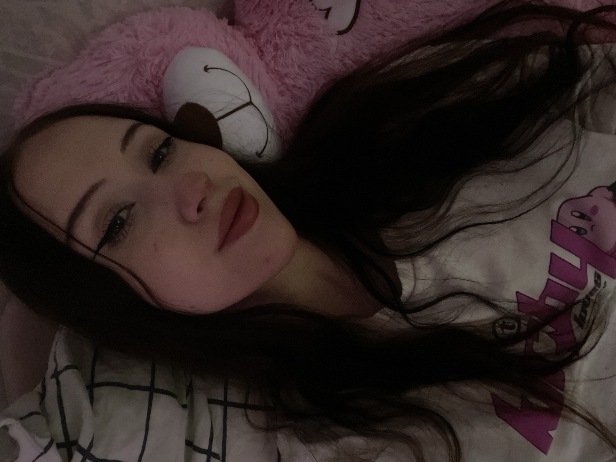 A young woman with long, dark hair is lying in bed, looking at the camera. She is wearing a white t-shirt with a pink graphic of Kirby on it. To her right is a large, fluffy pink teddy bear. The background consists of a white and black checkered duvet. The lighting is dim, casting a soft glow on the scene. This image was taken in Votkinsk, Russia.