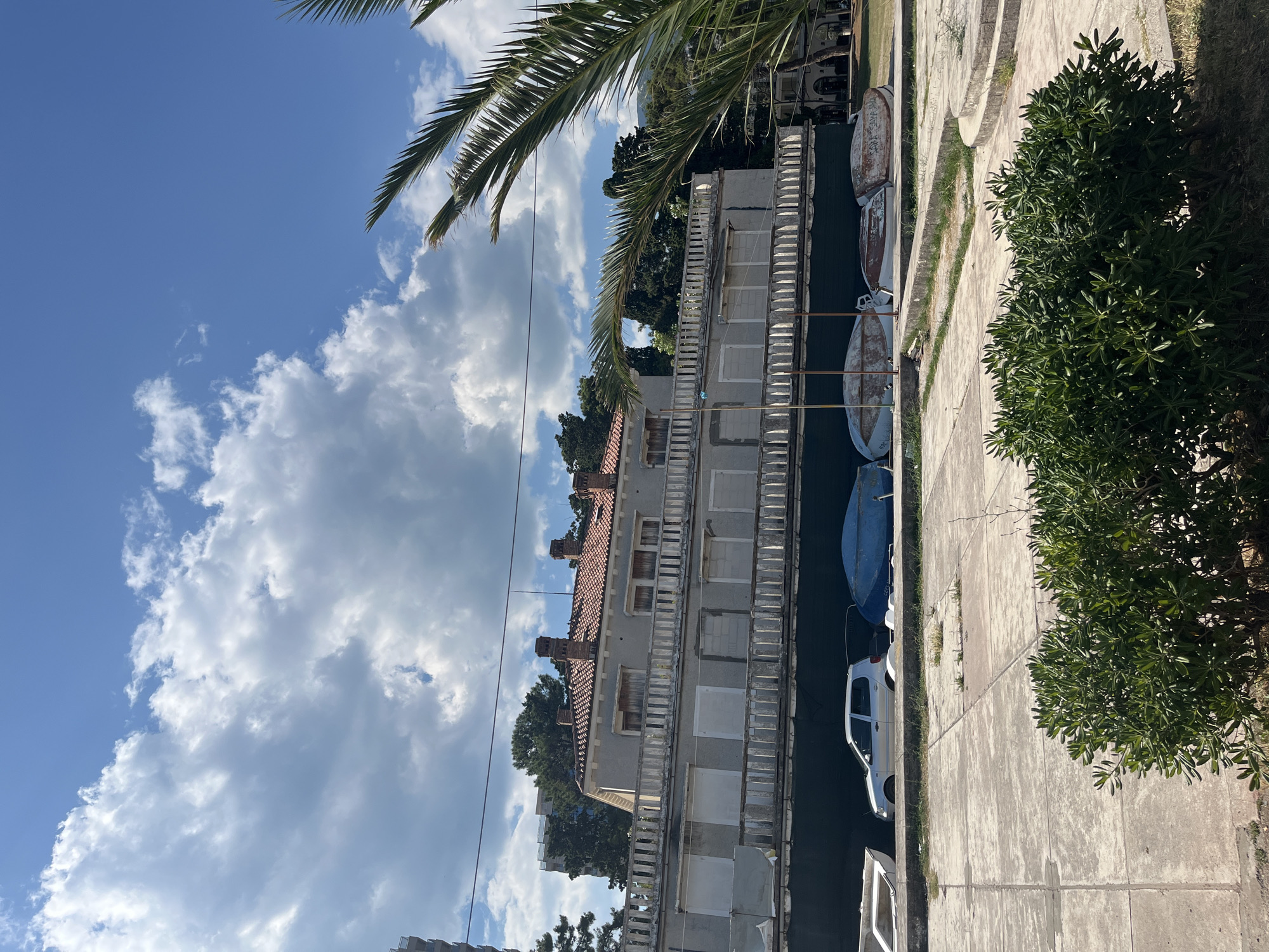 The image depicts a scene in Budva, Montenegro, likely during daytime under a clear blue sky with scattered white clouds. The perspective is from above, looking down at a narrow canal or waterway lined by a concrete walkway and a building.
The primary subjects are the waterway, the building, and several boats moored or resting in the water. The building appears to be a multi-story structure with a tiled roof, featuring a series of balconies with white railings. The facade is a light grey color, and some windows are visible, some boarded up. Trees and greenery surround the building, and a palm frond is visible in the upper left corner, suggesting a warm climate.
On the concrete walkway, a white car is parked. There are also several boats in the waterway, most of which are small and appear to be made of fiberglass. Some of the boats are a rusty brown color, while others are blue.
The overall impression is one of a peaceful, perhaps slightly weathered, coastal or canal-side environment. The lighting suggests it is a sunny day, casting shadows on the walkway. No people are visible in the frame. There is no discernible text in the image.