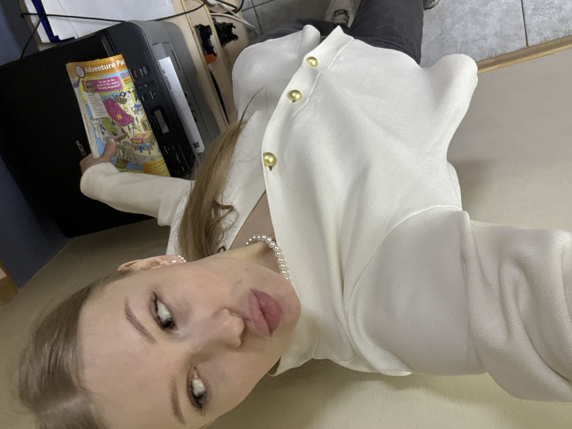A low-angle selfie depicts a woman indoors, possibly in Perm, Russia, given the potential location tag (though unconfirmed). She is fair-skinned with light blonde hair, wearing a white cardigan with gold buttons and a pearl necklace. She is holding a magazine titled "Adventure Park." A printer is visible in the background. The floor appears to be beige, and there are some cables and wires present.