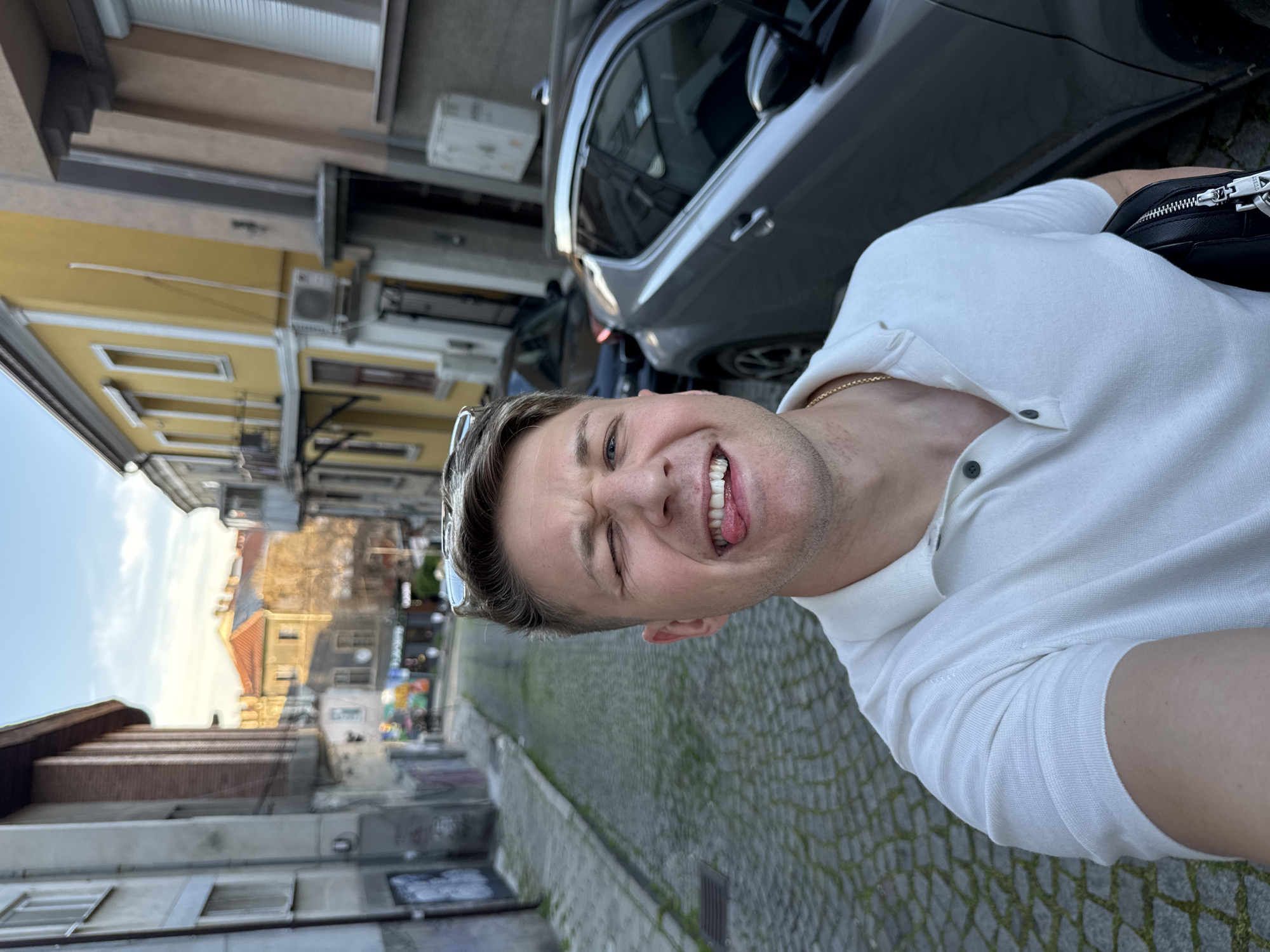 A selfie of a man with blond hair, wearing a white collared shirt and a gold chain, standing outdoors on a cobbled street in Plovdiv, Bulgaria. He has sunglasses on his head and is making a face, sticking out his tongue and winking. A black purse is visible. Cars are parked nearby on the street. Buildings with yellow facades line the street in the background.